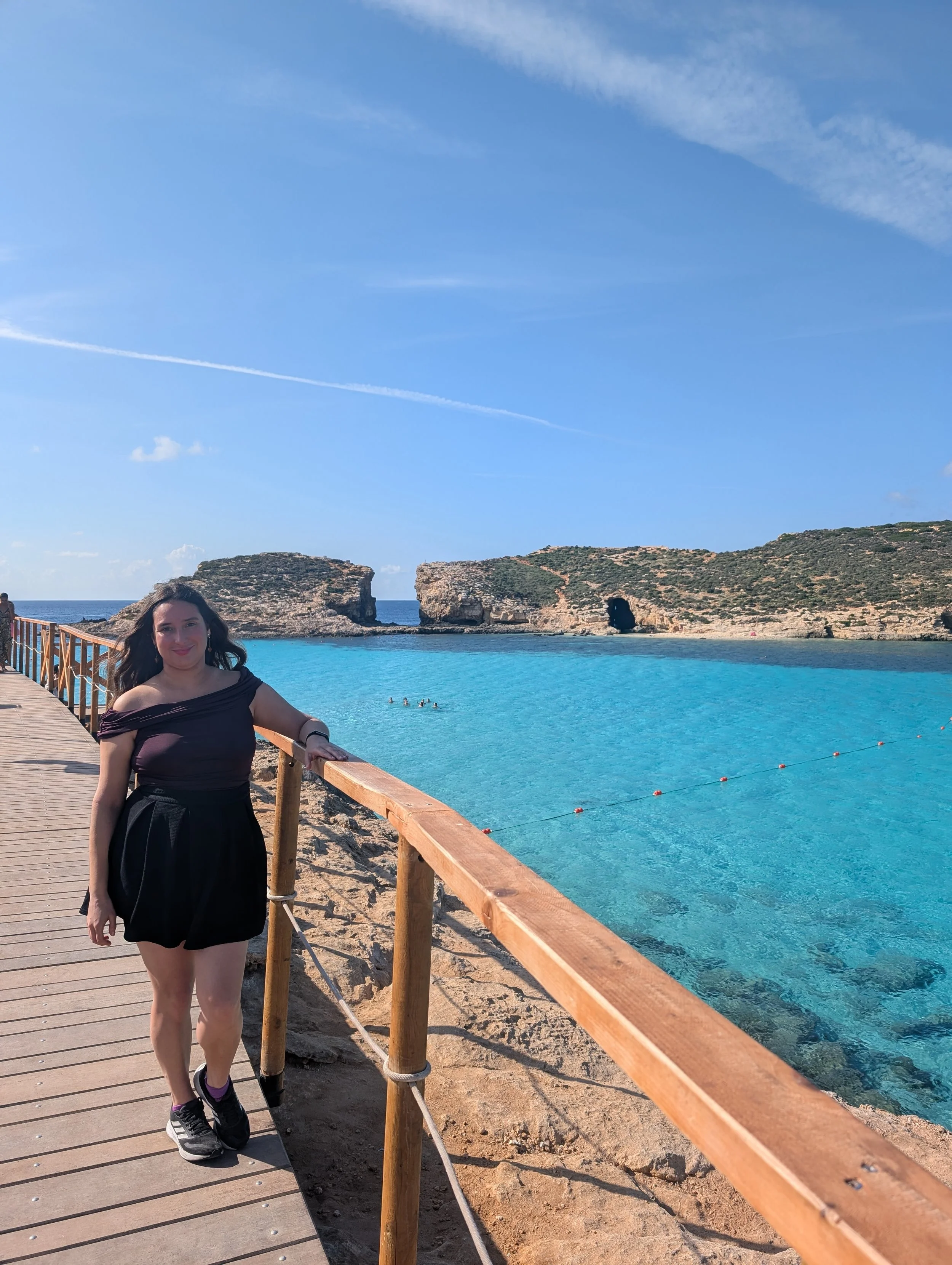 A woman with long dark hair, wearing a black off-shoulder top, shorts, and sneakers, standing on a wooden boardwalk beside an ocean with rocky cliffs and a clear blue sky in the background.