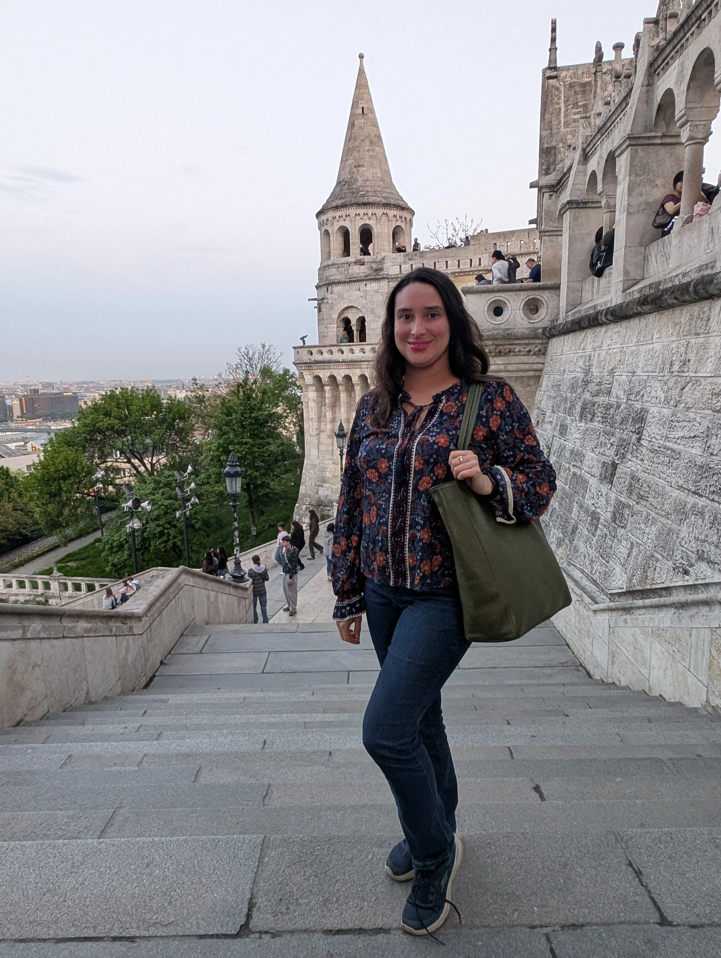 A woman with dark hair, wearing a floral jacket and jeans, standing on stone stairs in front of a historic castle with a turret and a cityscape in the background.