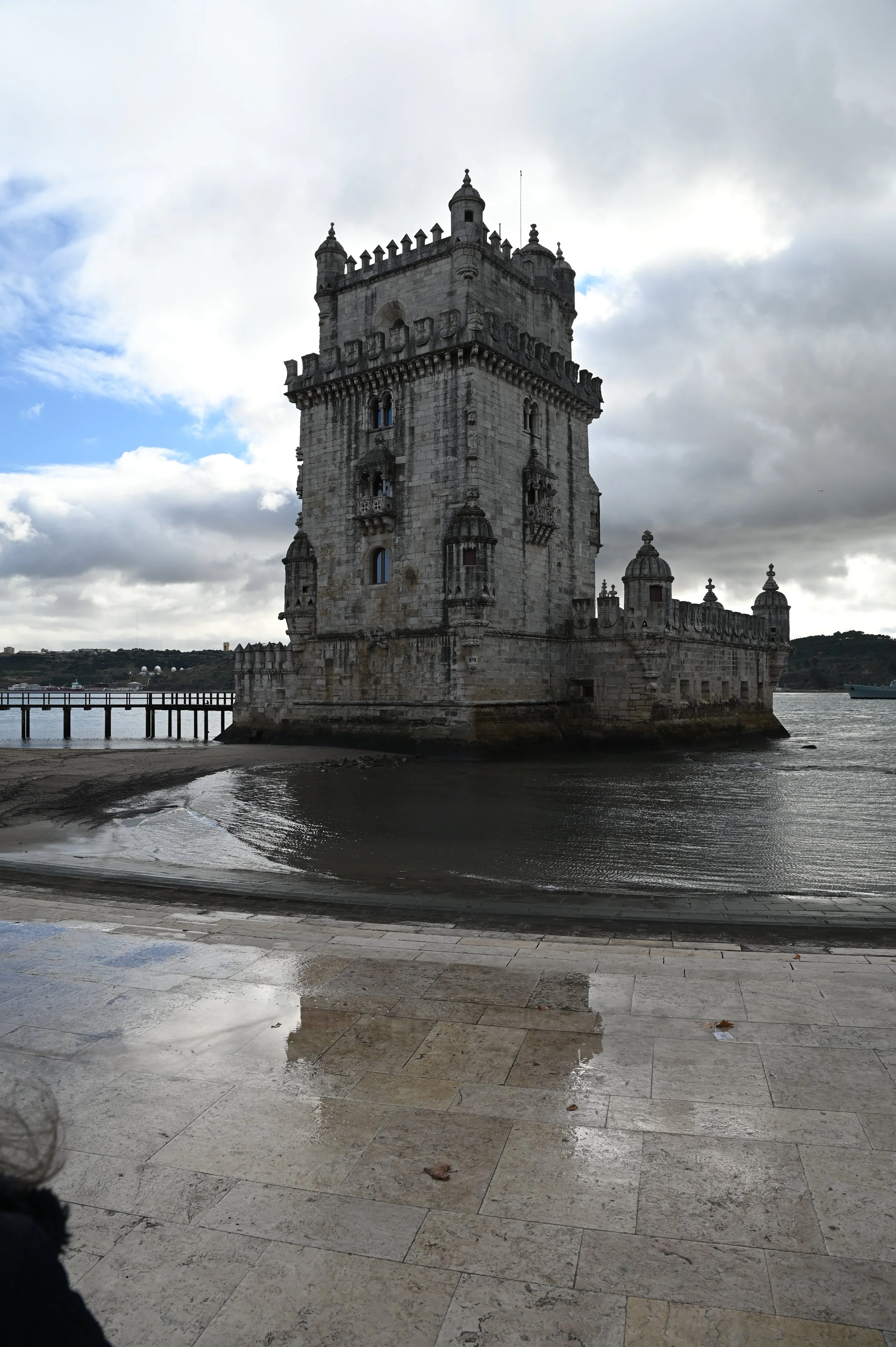 A historic stone tower with battlements and small turrets along the top, standing on the edge of a body of water with cloudy skies in the background.