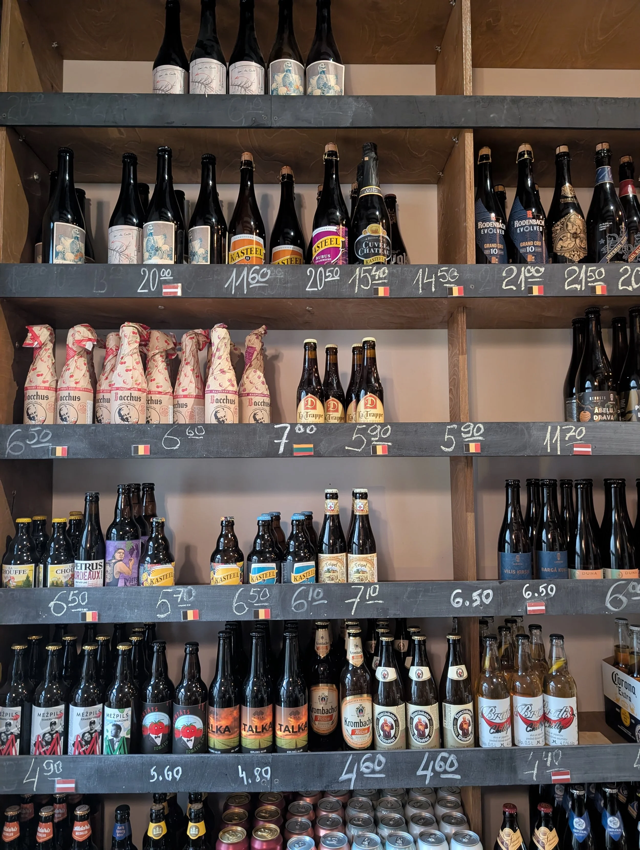 Shelf of assorted beer bottles and cans with price tags and flags, including Belgian and Lithuanian beers, in a liquor store.