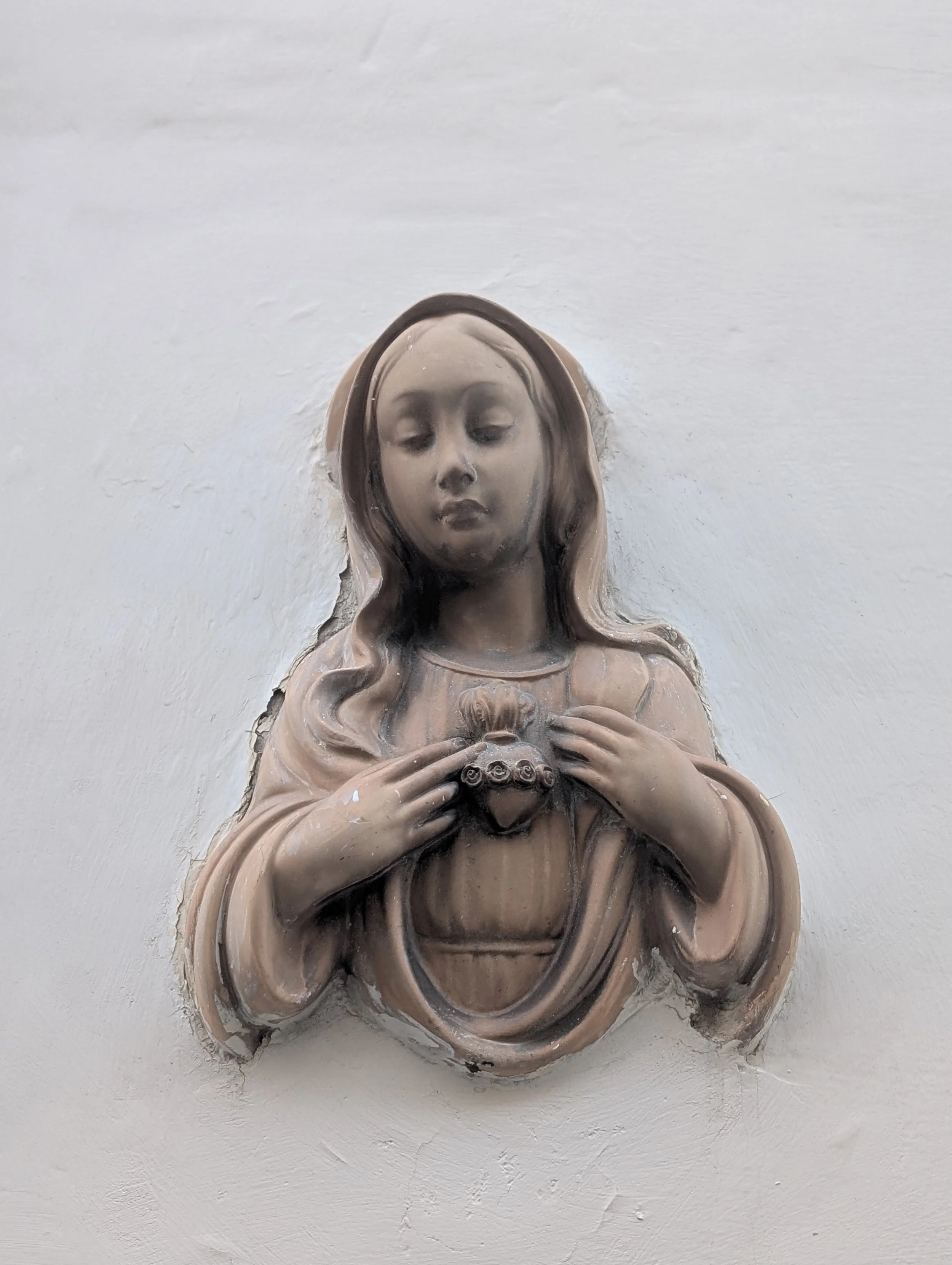 A bronze or brass statue of the Virgin Mary with hands clasped in prayer, mounted on a white wall.