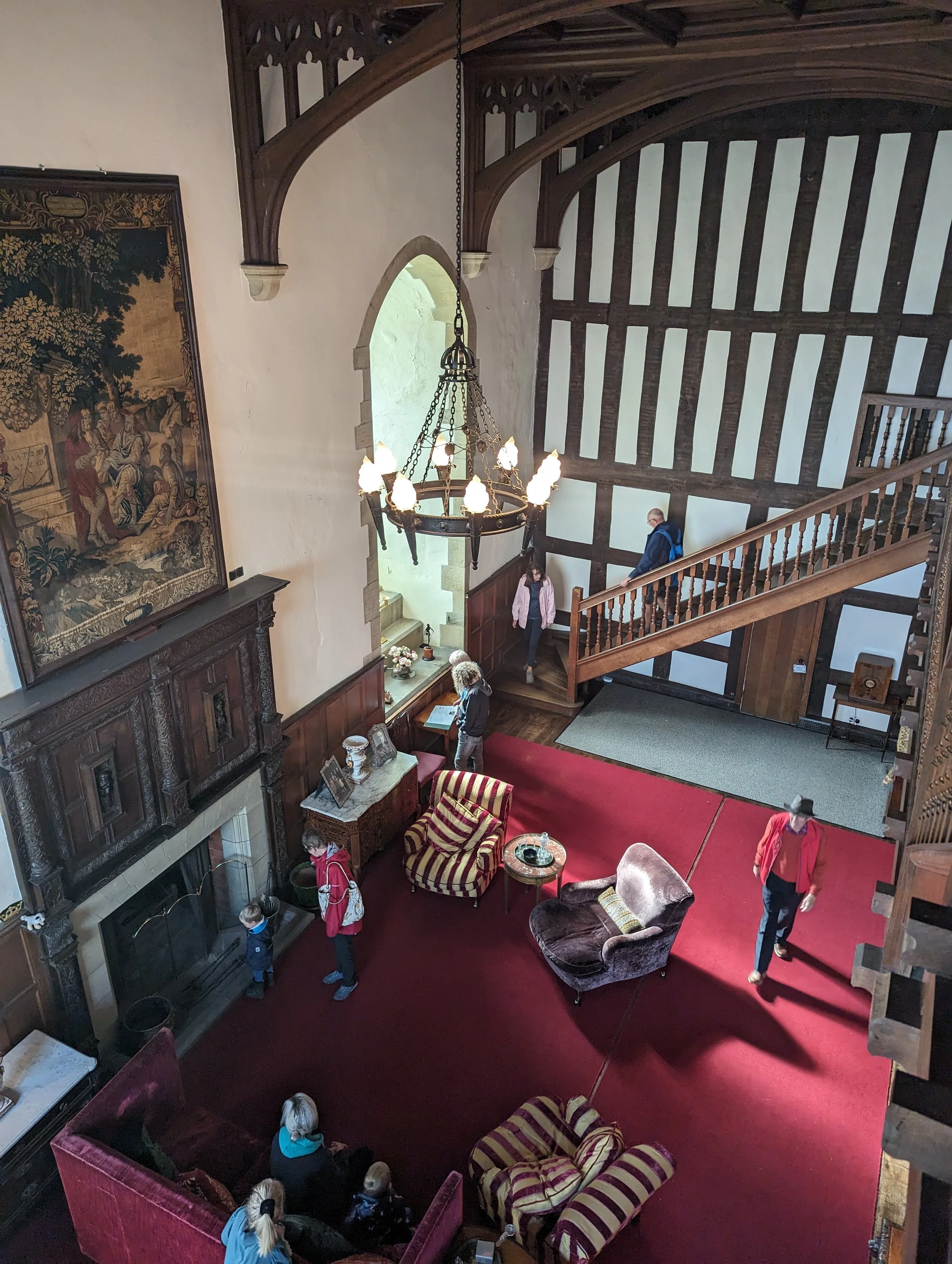 Interior view of a historic, wood-beamed hall with a large chandelier, a fireplace, antique furniture, and several people walking and sitting on sofas and armchairs.