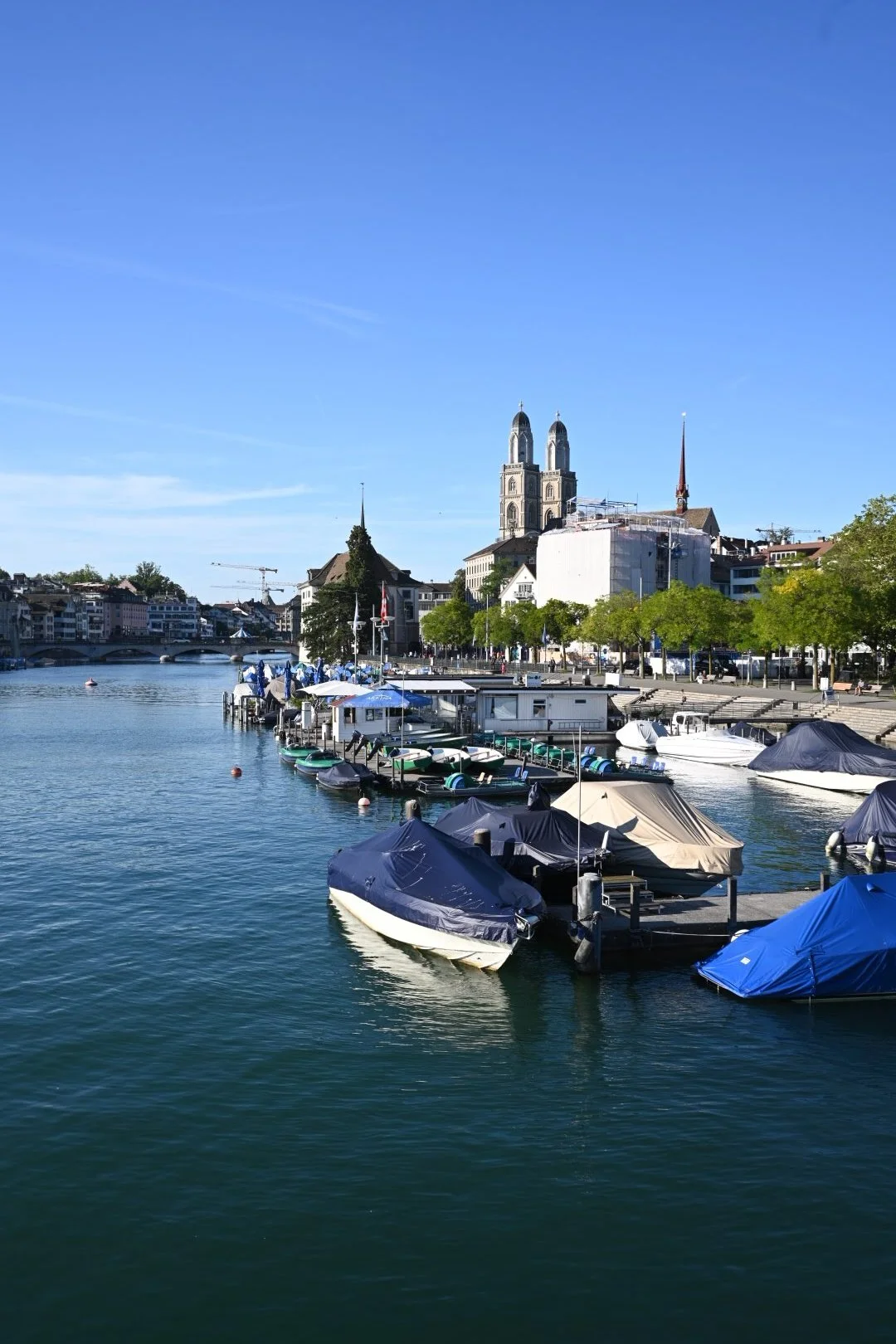 Boats docked along a river with a cityscape including historic buildings and church spires in the background under a clear blue sky.