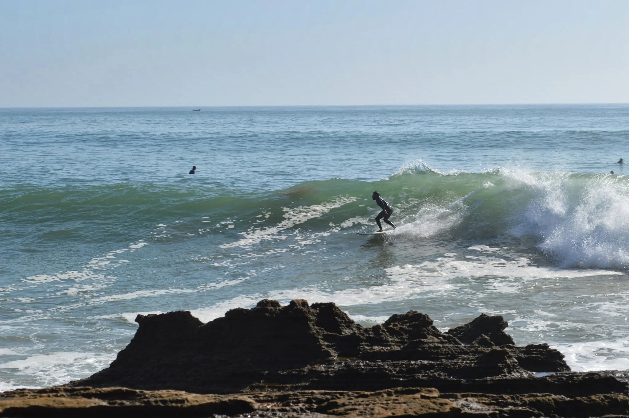 A beach scene with a surfer riding a wave and other surfers in the water, with rocky formations in the foreground and the open ocean stretching to the horizon.