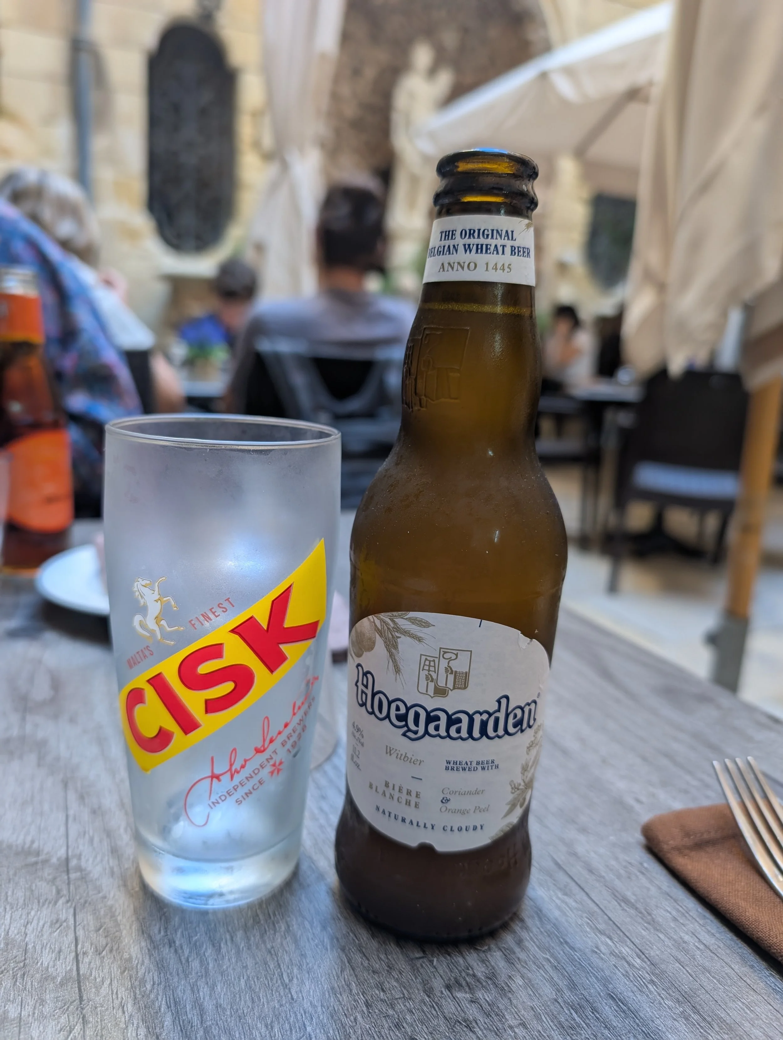 A beer bottle labeled 'Hoegaarden' on a gray wooden table next to a clear glass with a yellow and red logo.