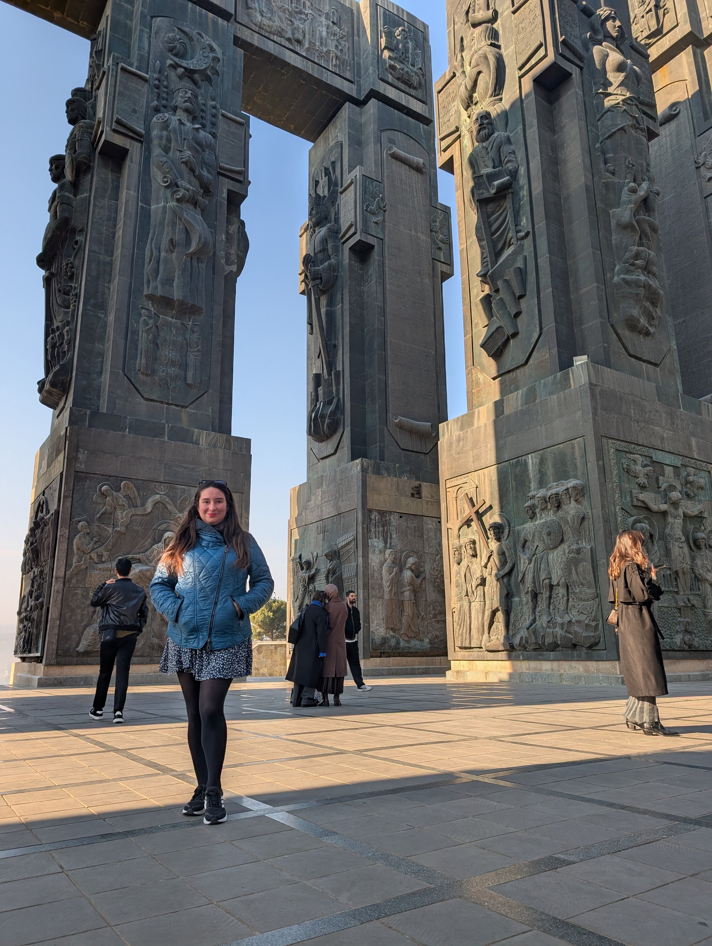 A woman in a blue jacket and black tights standing outside a large monument with carved figures and relief sculptures, with several other people in the background.