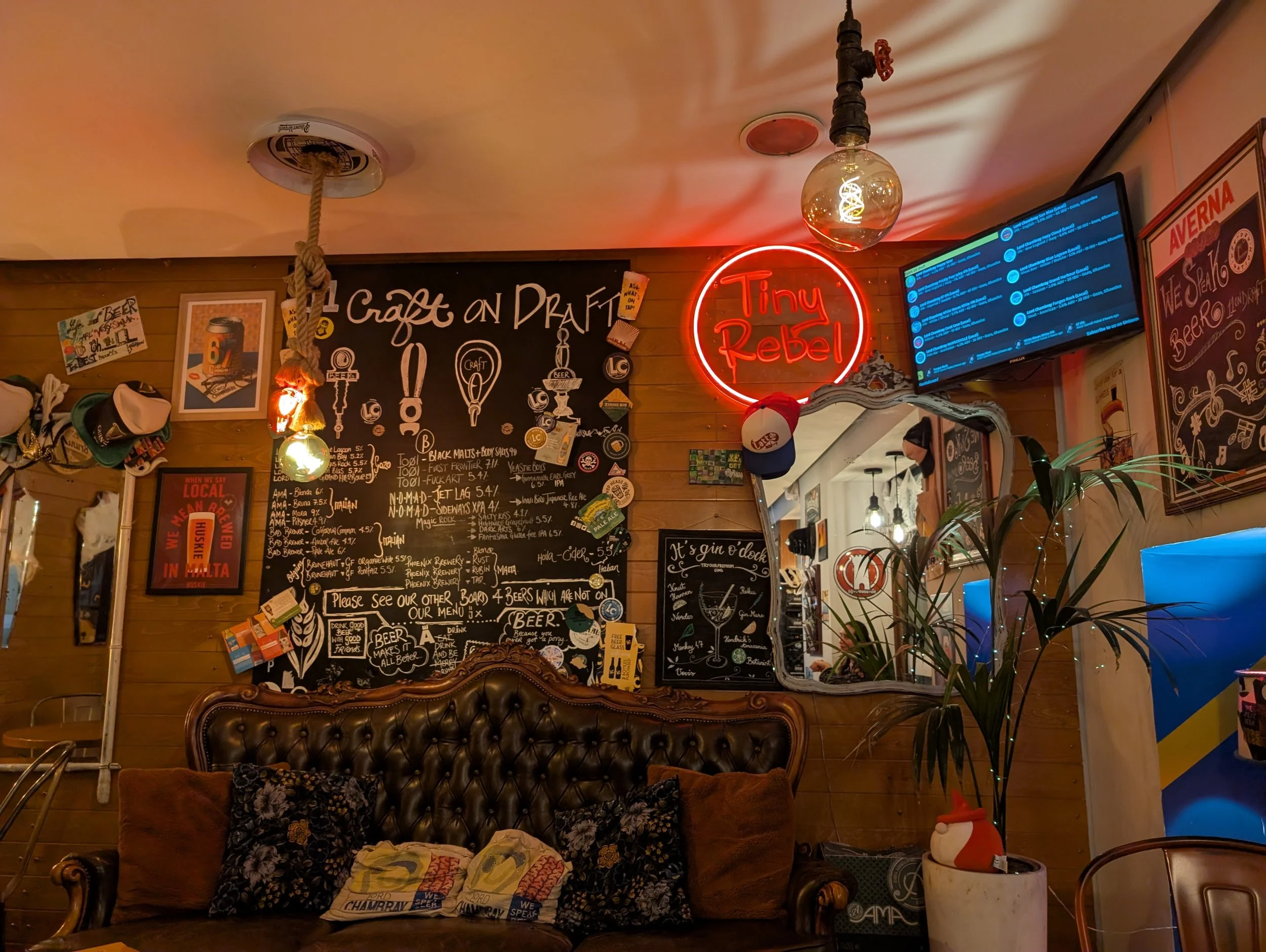 Interior of a bar or restaurant with a blackboard menu, neon sign saying 'Tiny Rebel,' and various decorations on the walls. There are hanging light bulbs, a TV screen, and a plant in a white pot.