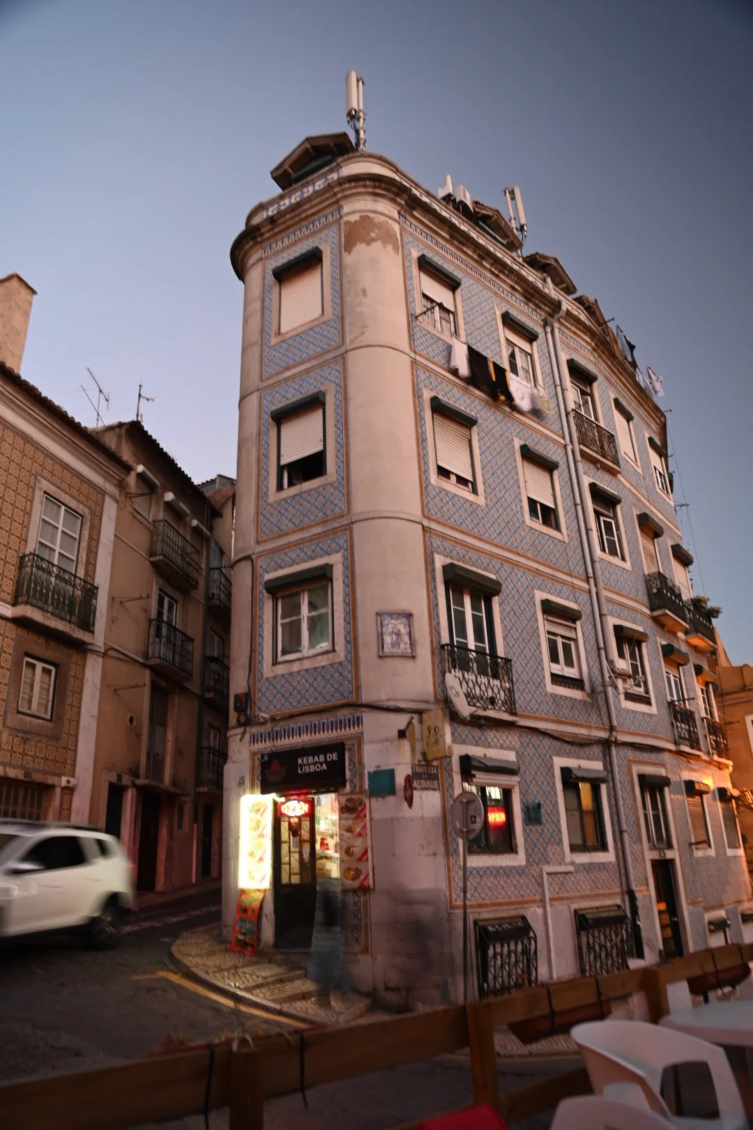 A multi-story building with traditional Portuguese tile facade, laundry hanging from windows, and a small kebab shop at the corner during dusk.