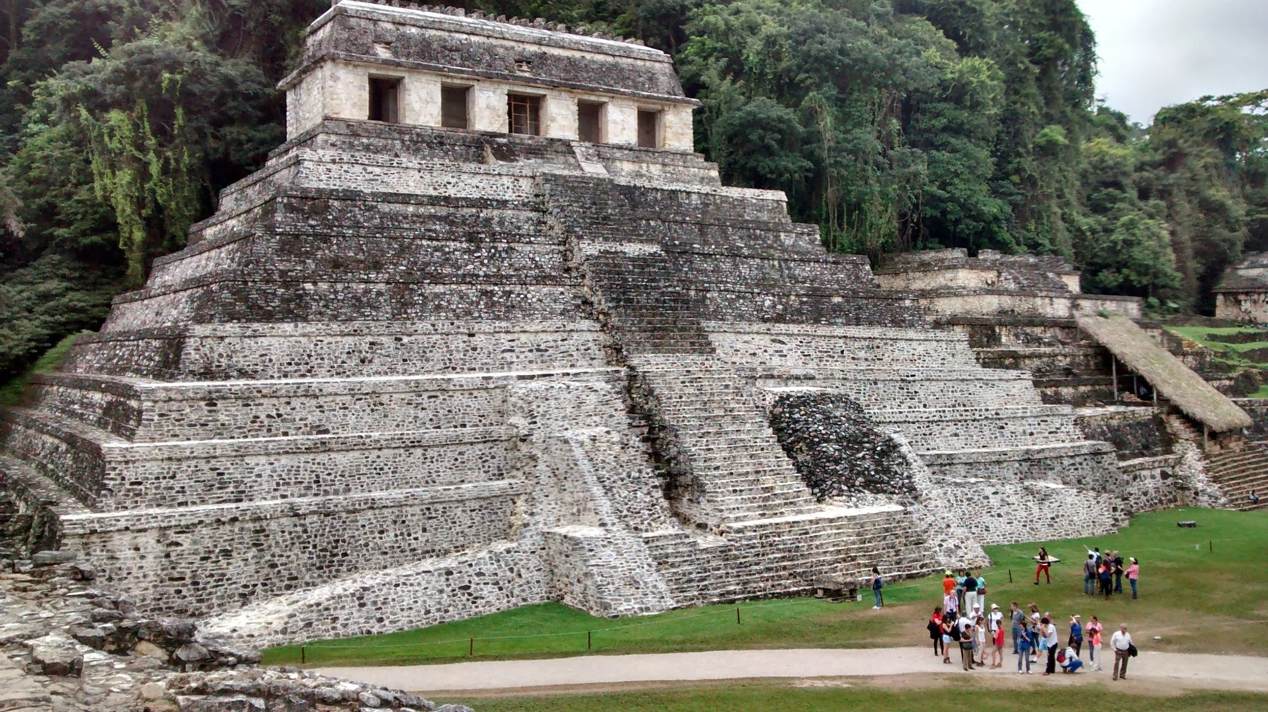 Ancient Mayan pyramid with multiple levels made of stone and surrounded by greenery, with groups of tourists at the base.