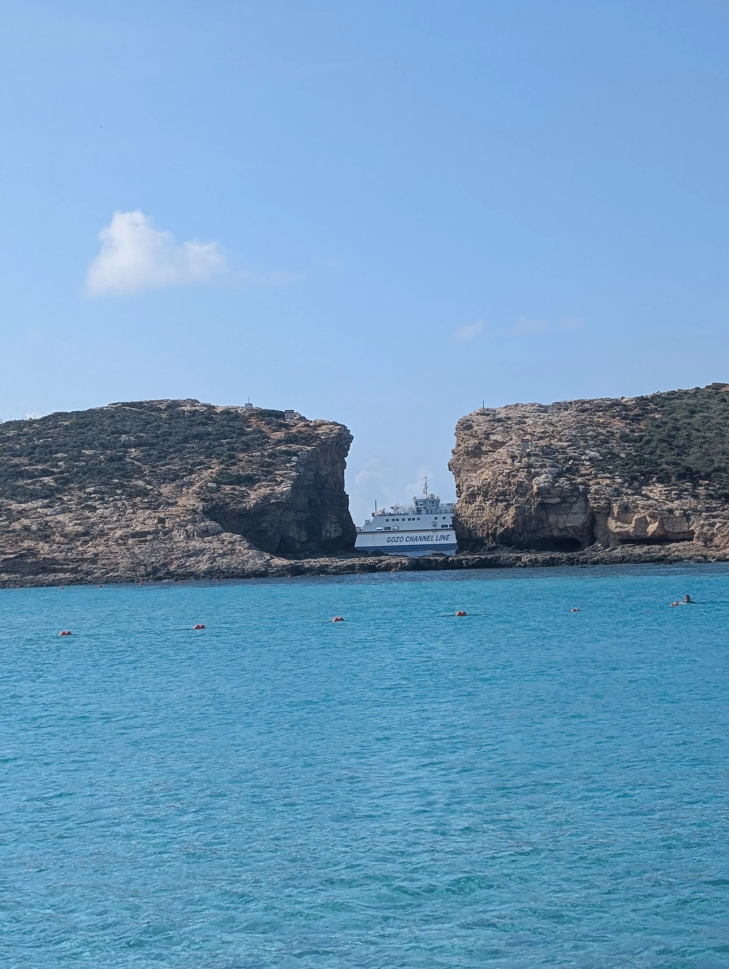 A scenic view of ocean water with two large rocky cliffs and a ferry boat between them, under a clear blue sky with a few clouds.
