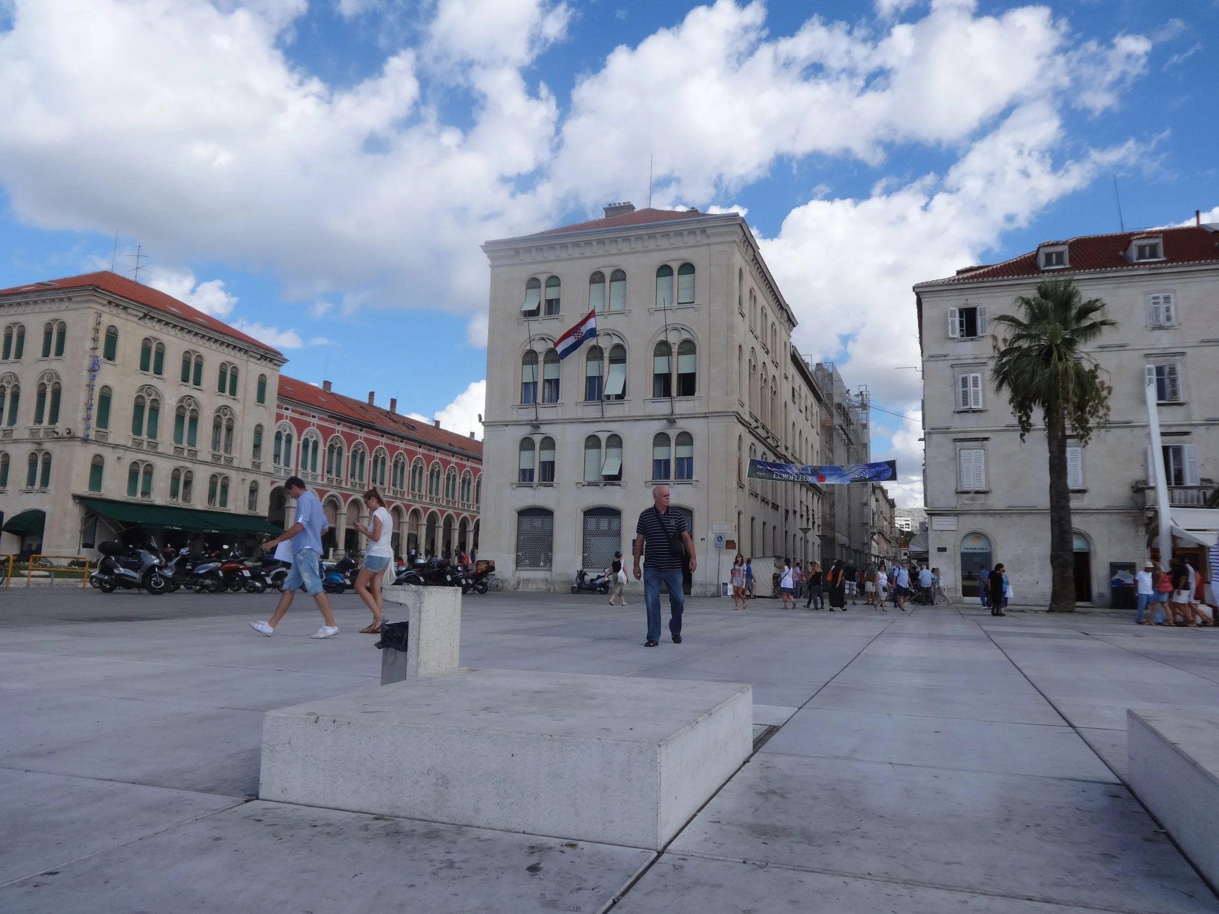 A busy city square with people walking, motorcycles parked along the street, and historic white buildings with red roofs under a partly cloudy sky. A Croatian flag is visible on a building.