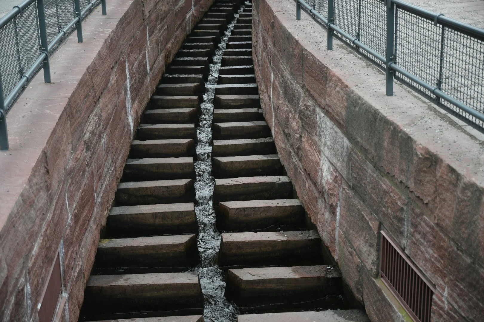 A narrow stone-lined water channel or sluice with wooden slats across the top, allowing water to flow down the center. The channel is bordered by sidewalks with metal railings.
