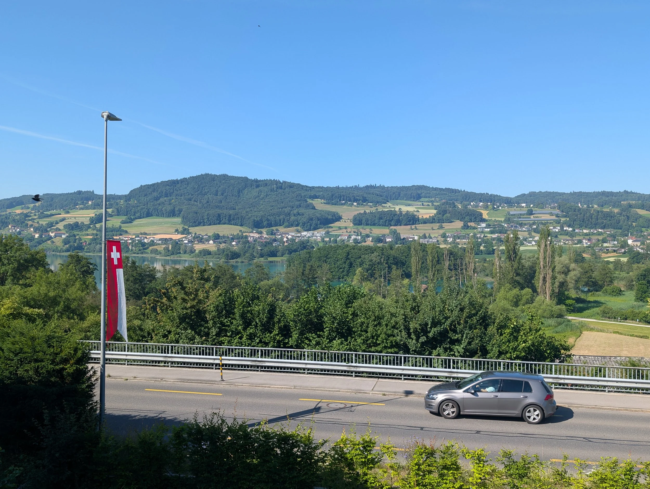 A scenic view of rolling green hills and a small town beneath a clear blue sky, with a Swiss flag partially visible in the foreground, a road with a parked car, and a streetlamp.