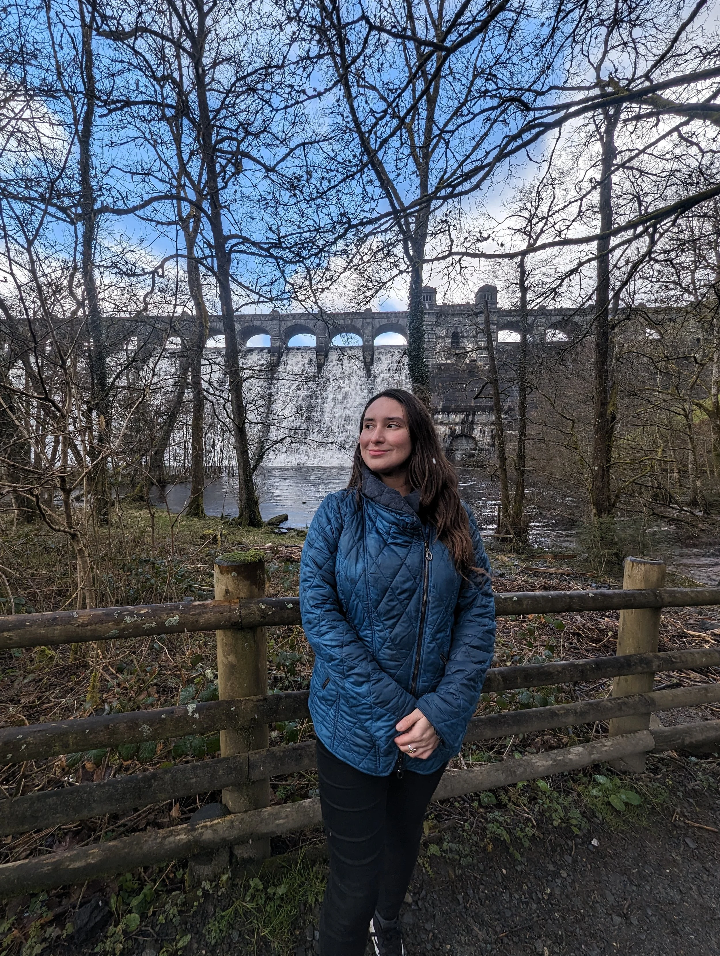 A woman with long dark hair, wearing a blue jacket and black pants, standing outdoors in front of a wooden fence. In the background, a dam with multiple arches and an old stone bridge can be seen, surrounded by leafless trees and a partly cloudy sky.