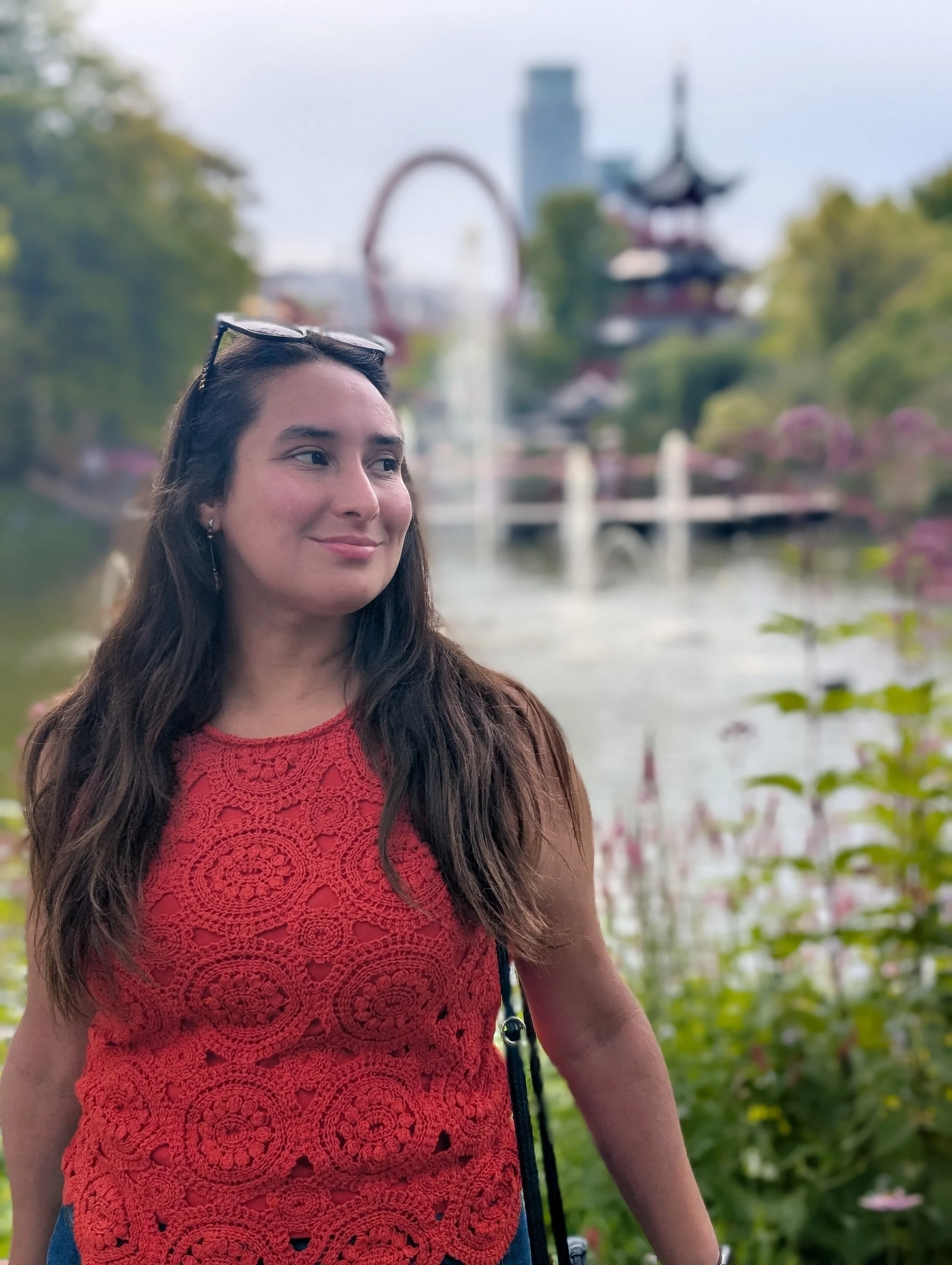 A woman with long dark hair and sunglasses resting on her head wearing a red lace sleeveless top, standing outdoors near a pond or lake in front of a fountain, trees, and a traditional Asian-style pavilion with pagoda roof.