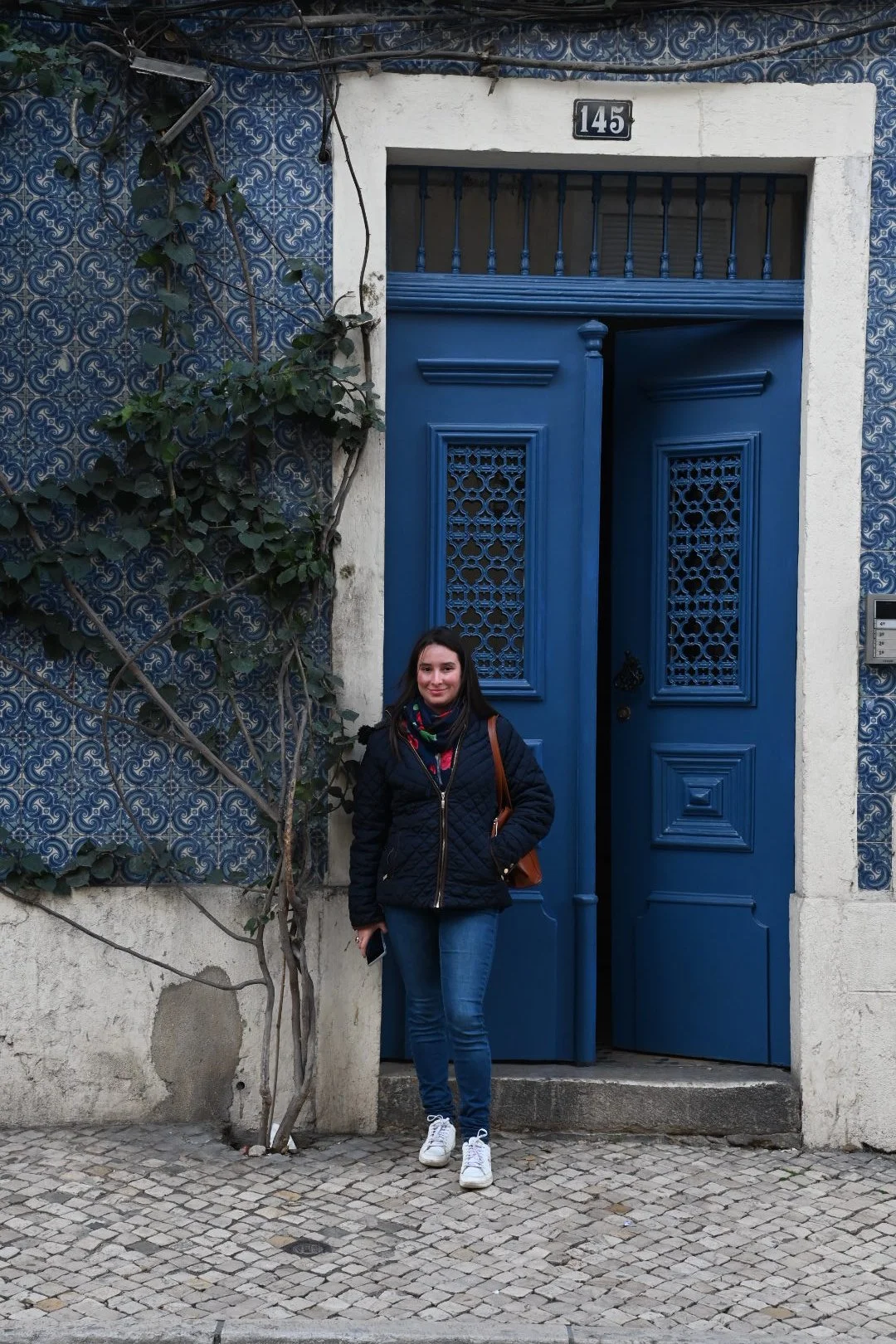 A woman in casual attire standing in front of a vibrant blue door with intricate grillwork, surrounded by a white wall and decorative blue tiles, on a cobblestone street.