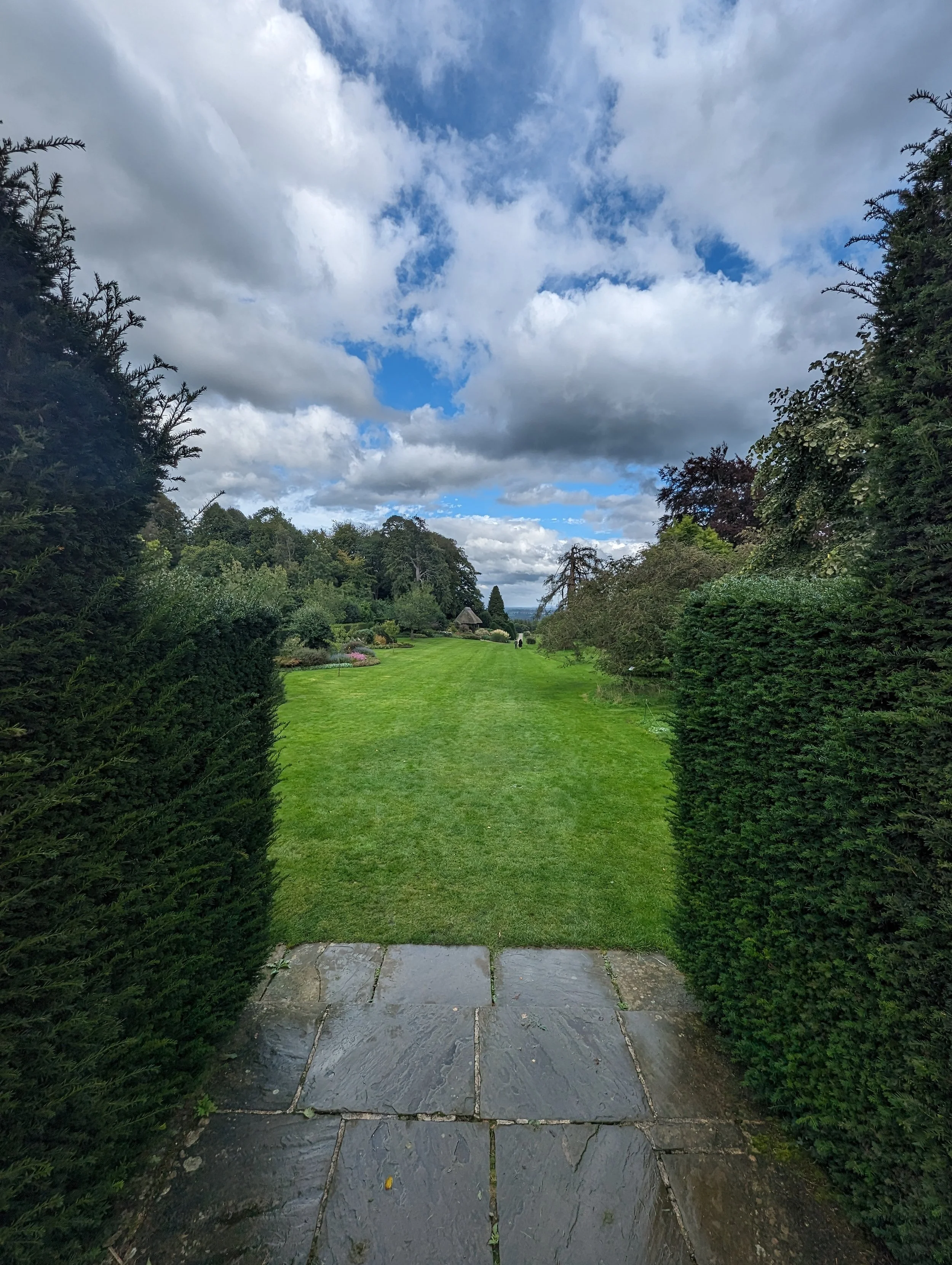 View of a garden with a stone pathway leading to a lush green lawn, surrounded by bushes and trees, under a partly cloudy sky.