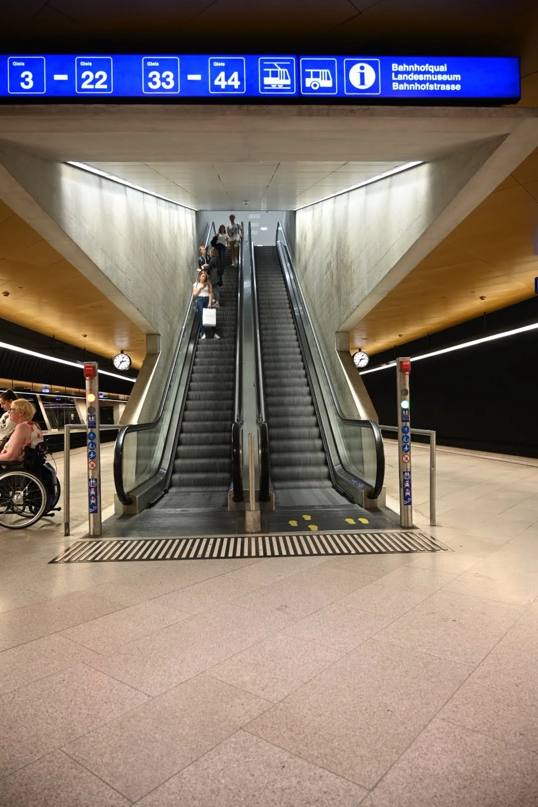Escalator in a train station with people and a person in a wheelchair, digital signs with information, and clocks on the walls.