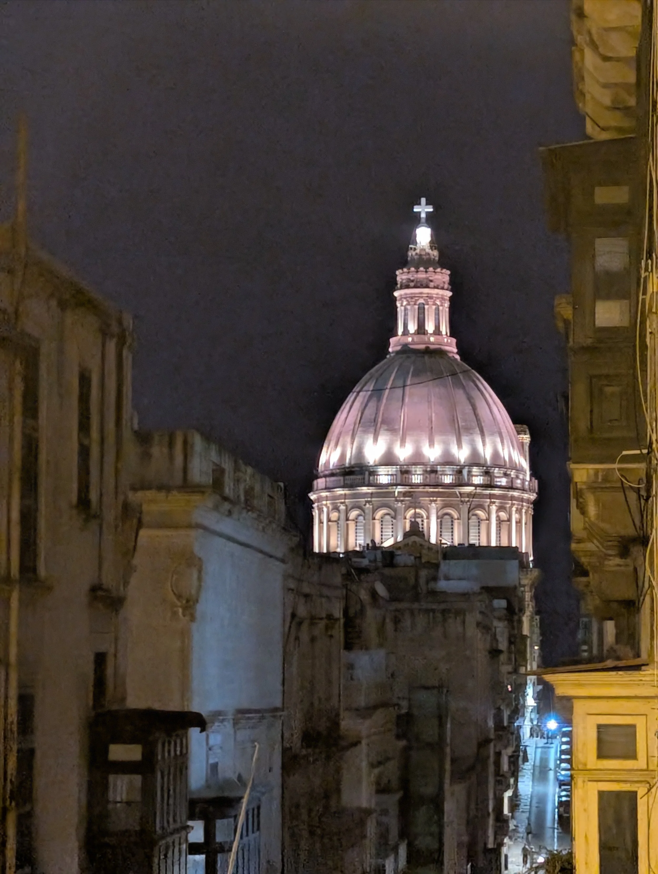 Night view of a city street with historical buildings on either side and a church or cathedral with a lit dome and cross in the background.