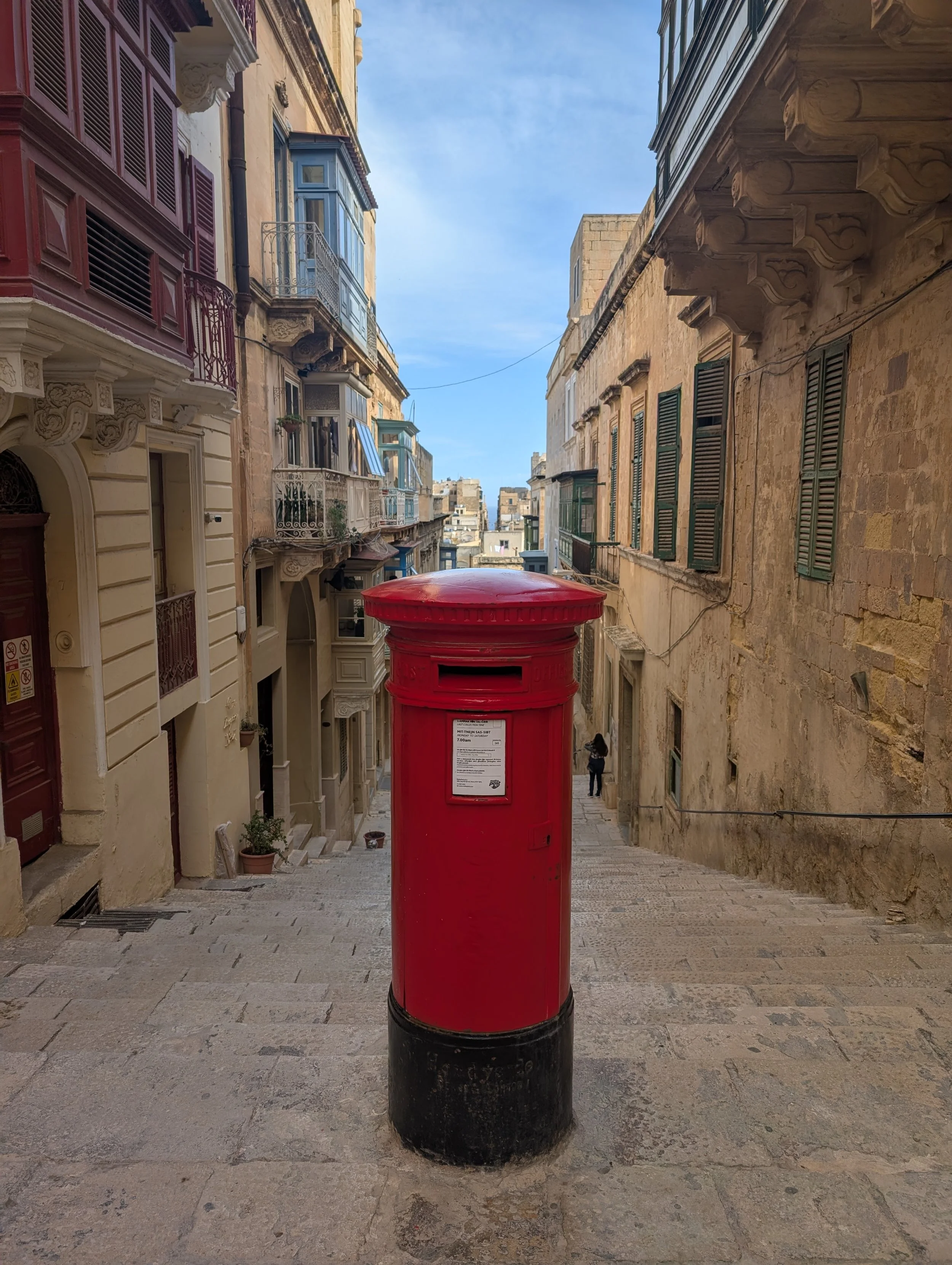A red mailbox located on a stone-paved street in a historic European city with old stone and stucco buildings with balconies.