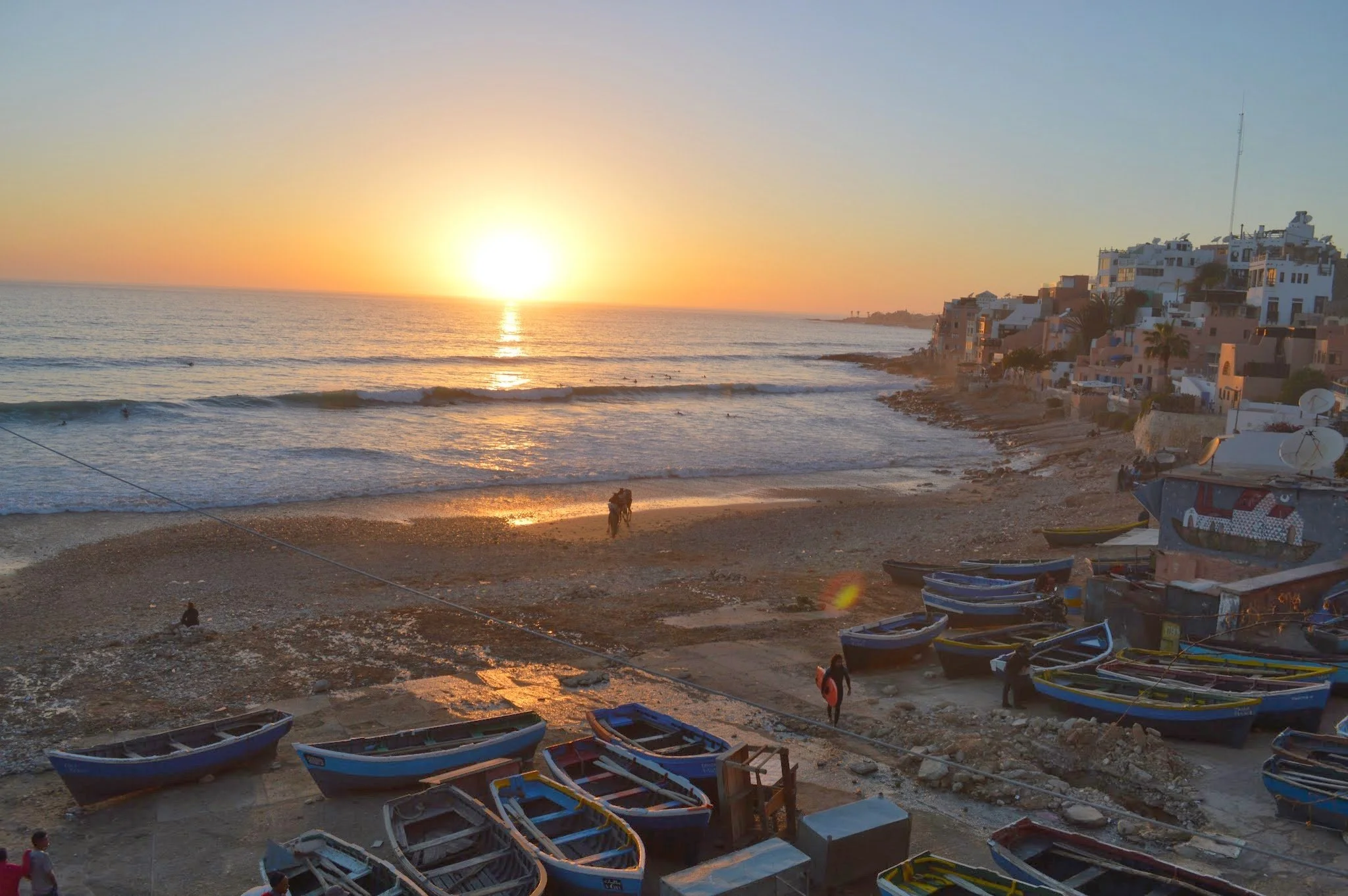 Sunset over the ocean with a beach and colourful houses on the right, several small boats on the shore, and people walking near the water.