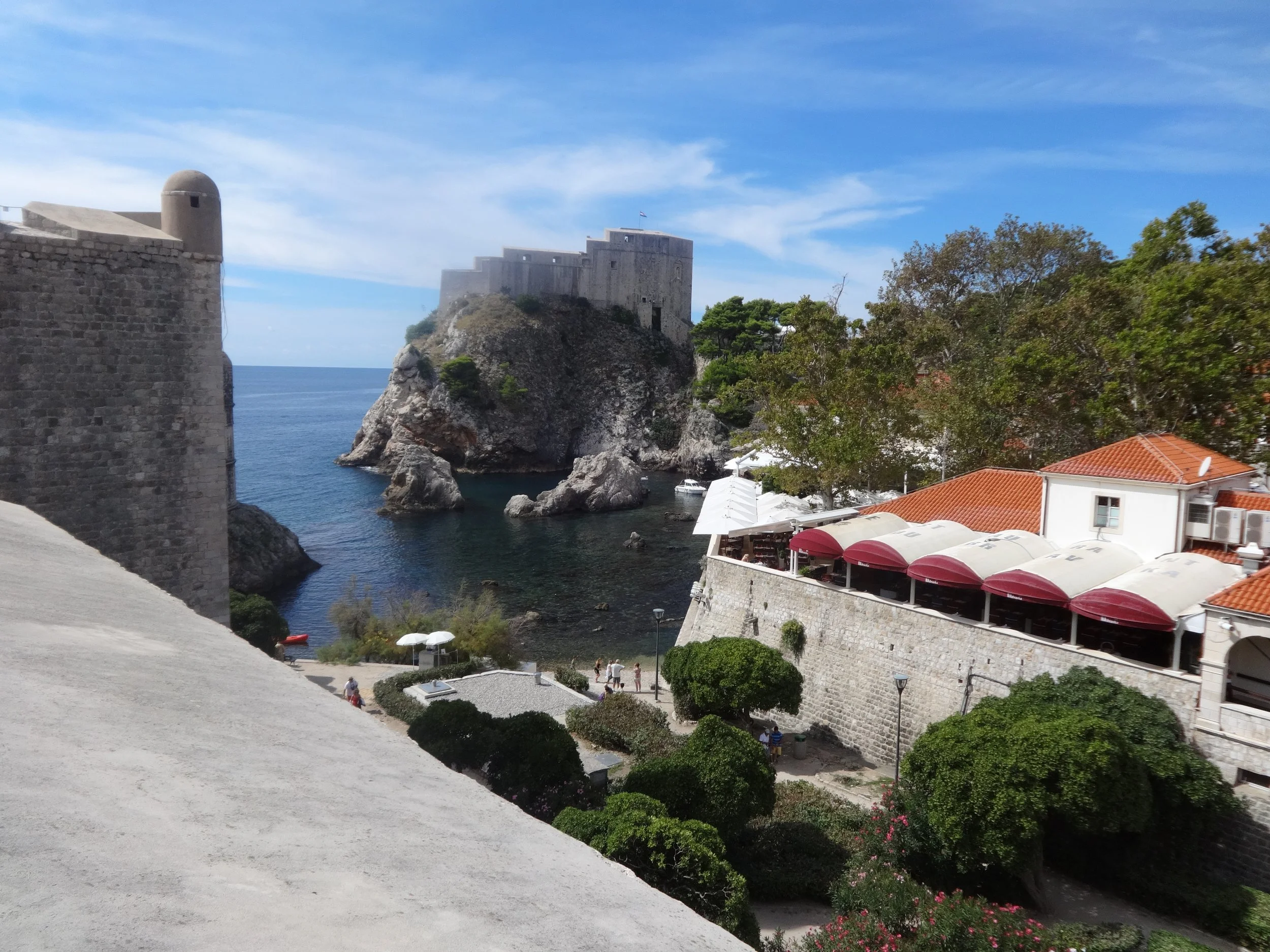 Fortress on a rocky cliff by the sea, with trees, boats, and a building with red-roofed terraces in the foreground, under a partly cloudy sky.