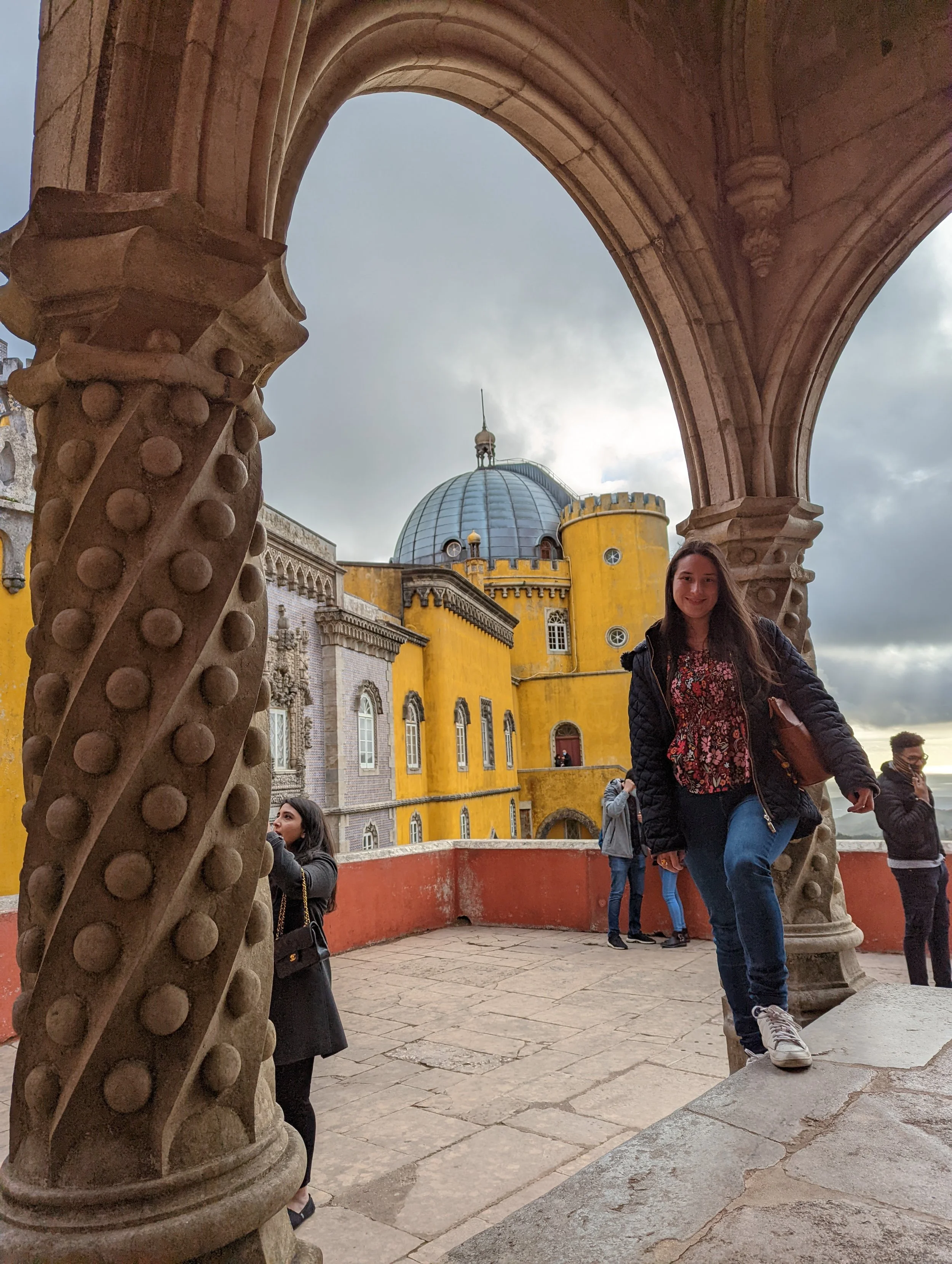 People standing and walking under a stone arch near a yellow and blue historic building with a dome, during cloudy weather.