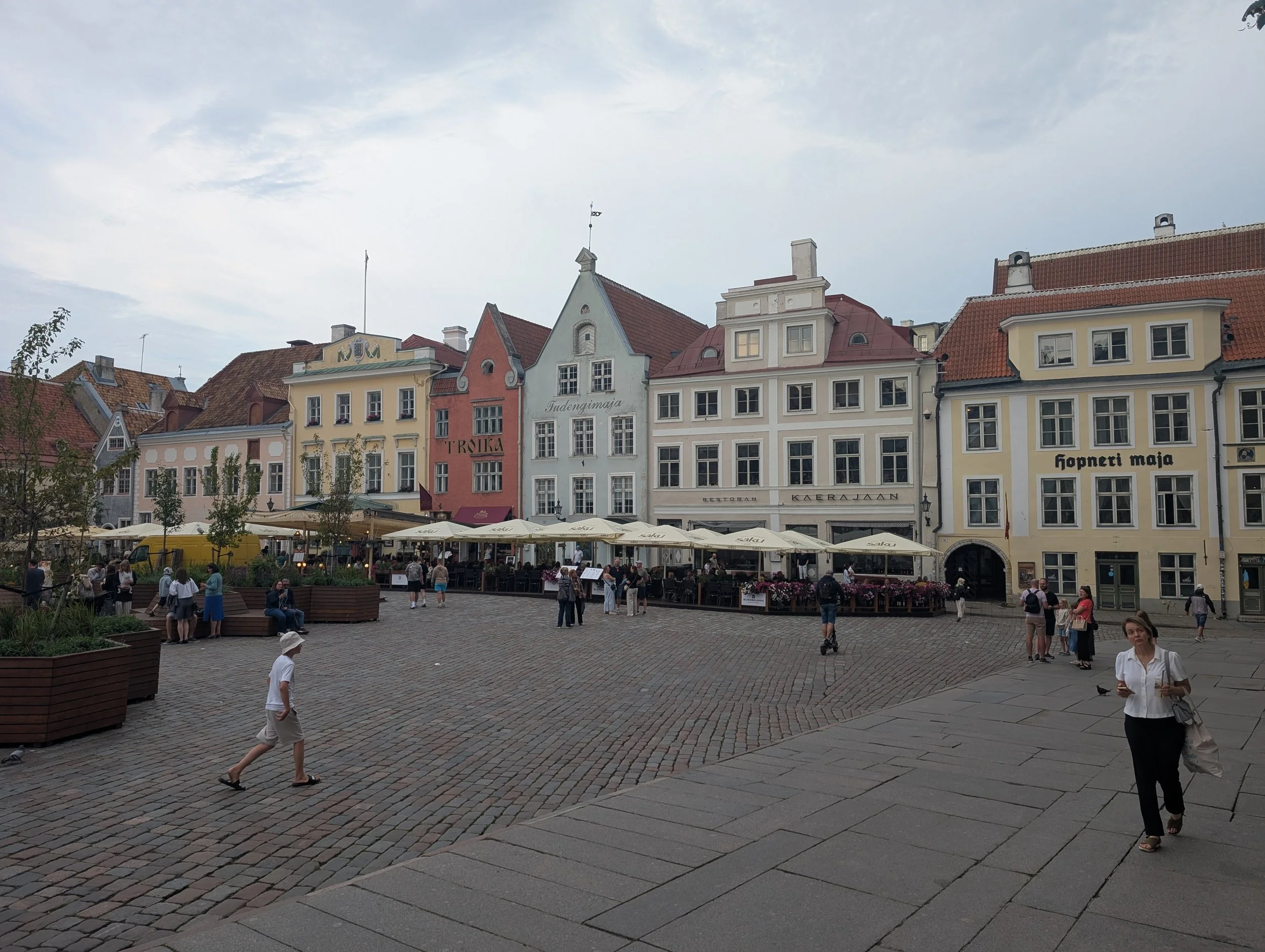 A European town square with colorful historic buildings, outdoor cafes with umbrellas, and people walking and sitting on benches.