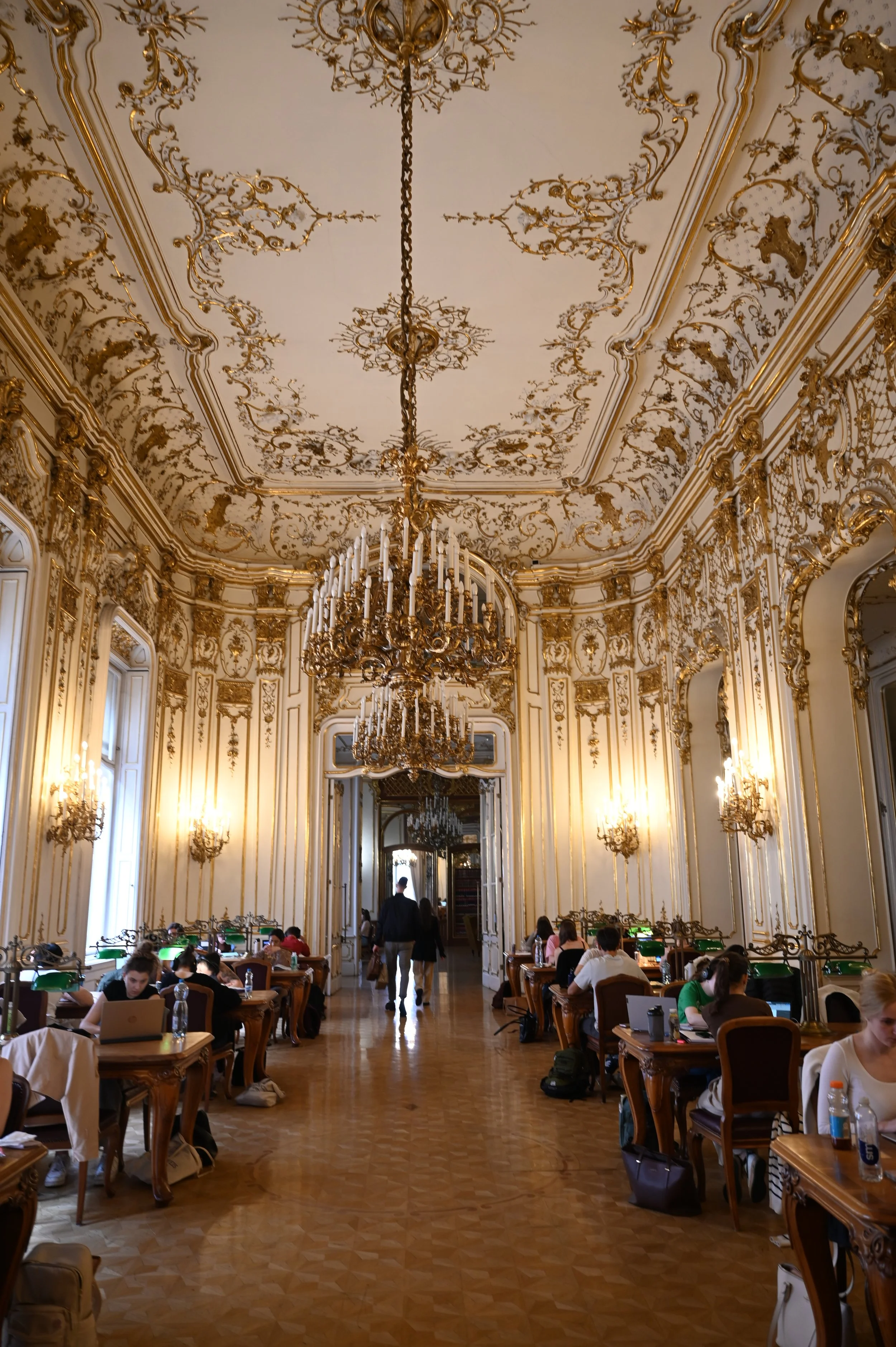 A grand, ornate room with high ceilings decorated with gold embellishments, large chandeliers, and tall windows. People are sitting at desks working on computers, and others are walking or standing in the hallway. The room has a classic, opulent styl