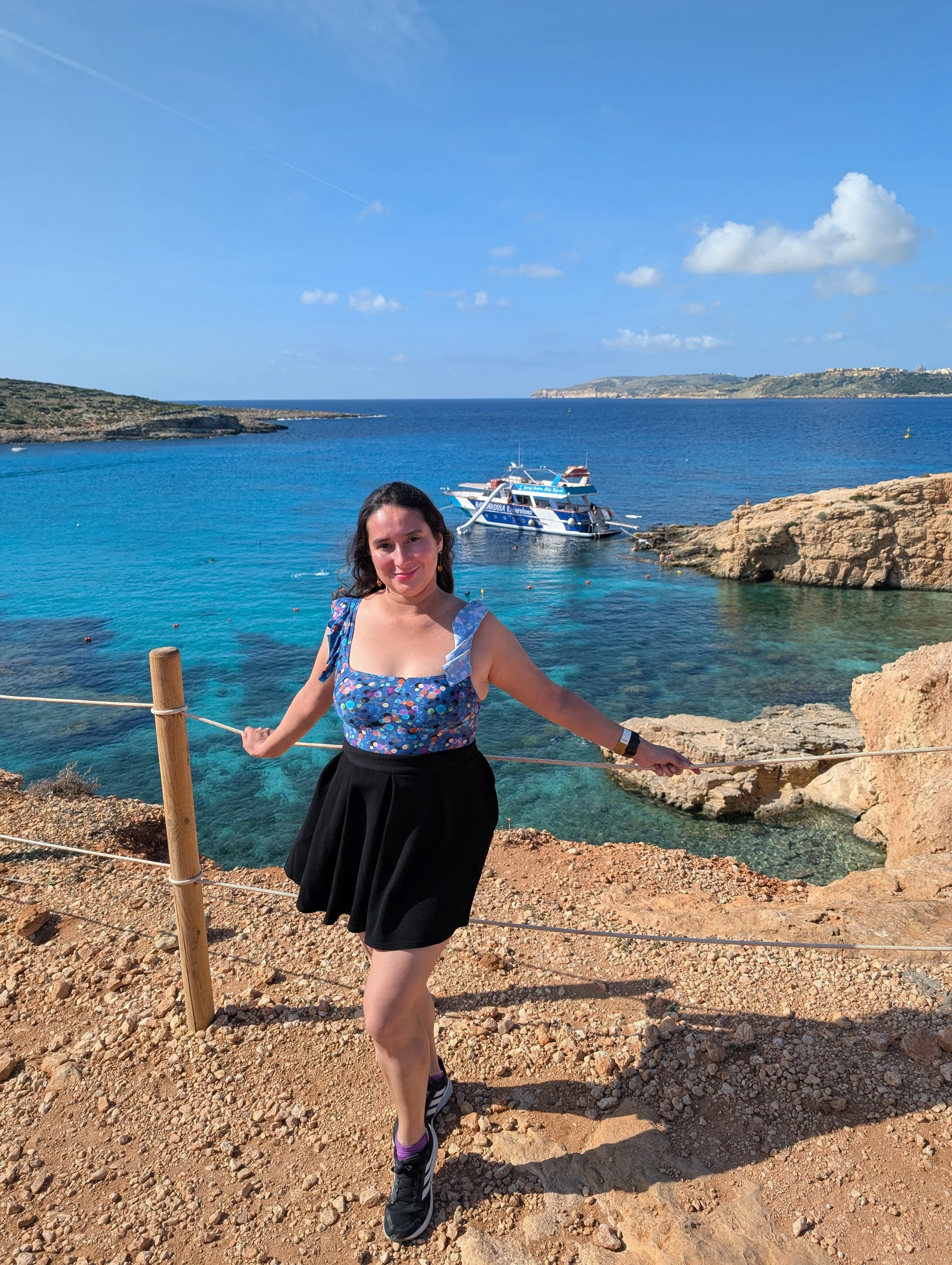 A woman in a blue floral top and black skirt standing on rocky ground near the coast, with blue water, a boat, and distant cliffs under a partly cloudy sky in the background.
