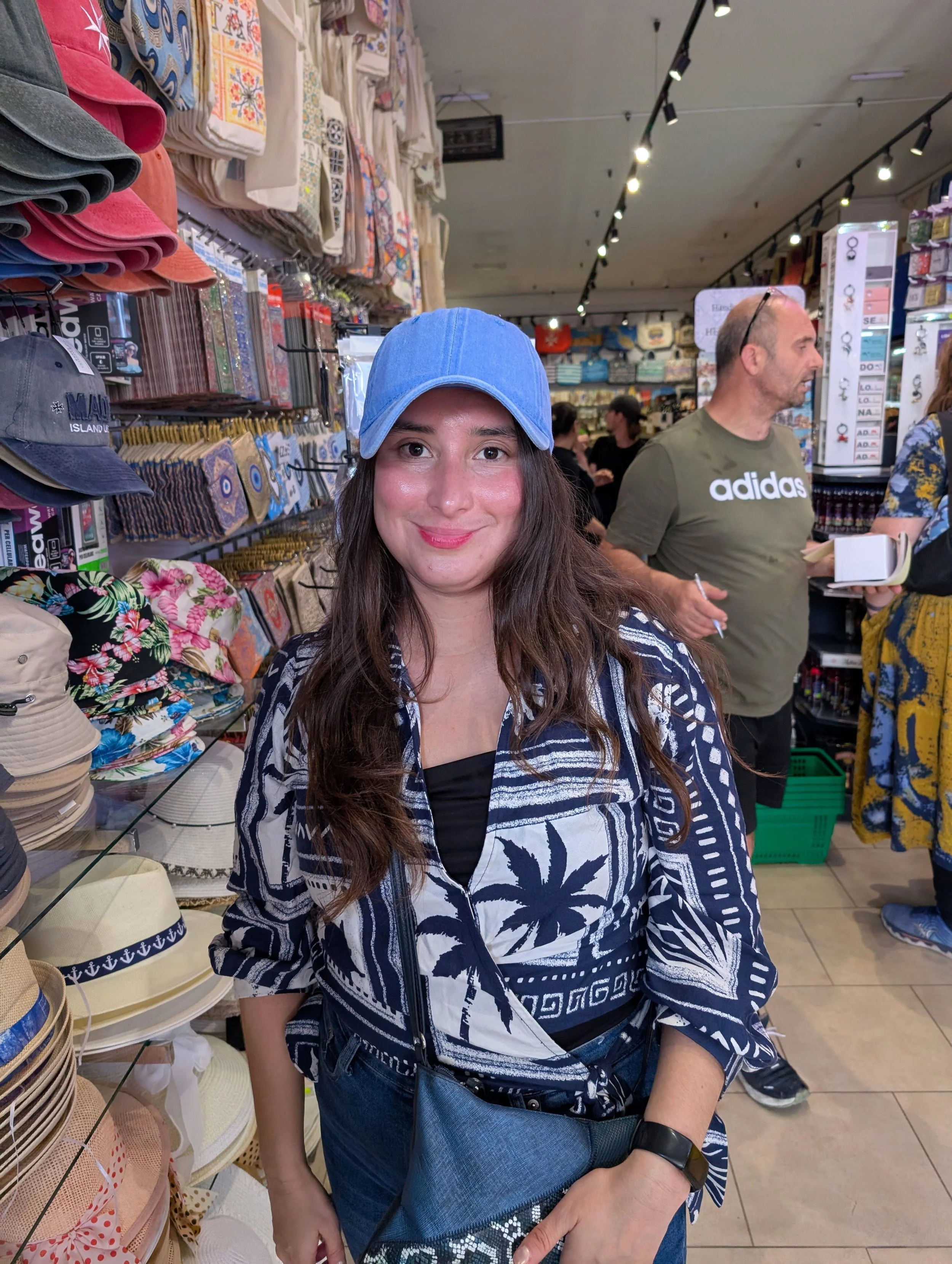 A young woman with long brown hair, wearing a blue cap, a black top, and a patterned blue-and-white jacket, is shopping in a store aisle filled with hats, bags, and accessories. She is smiling and holding a black-and-white woven bag.
