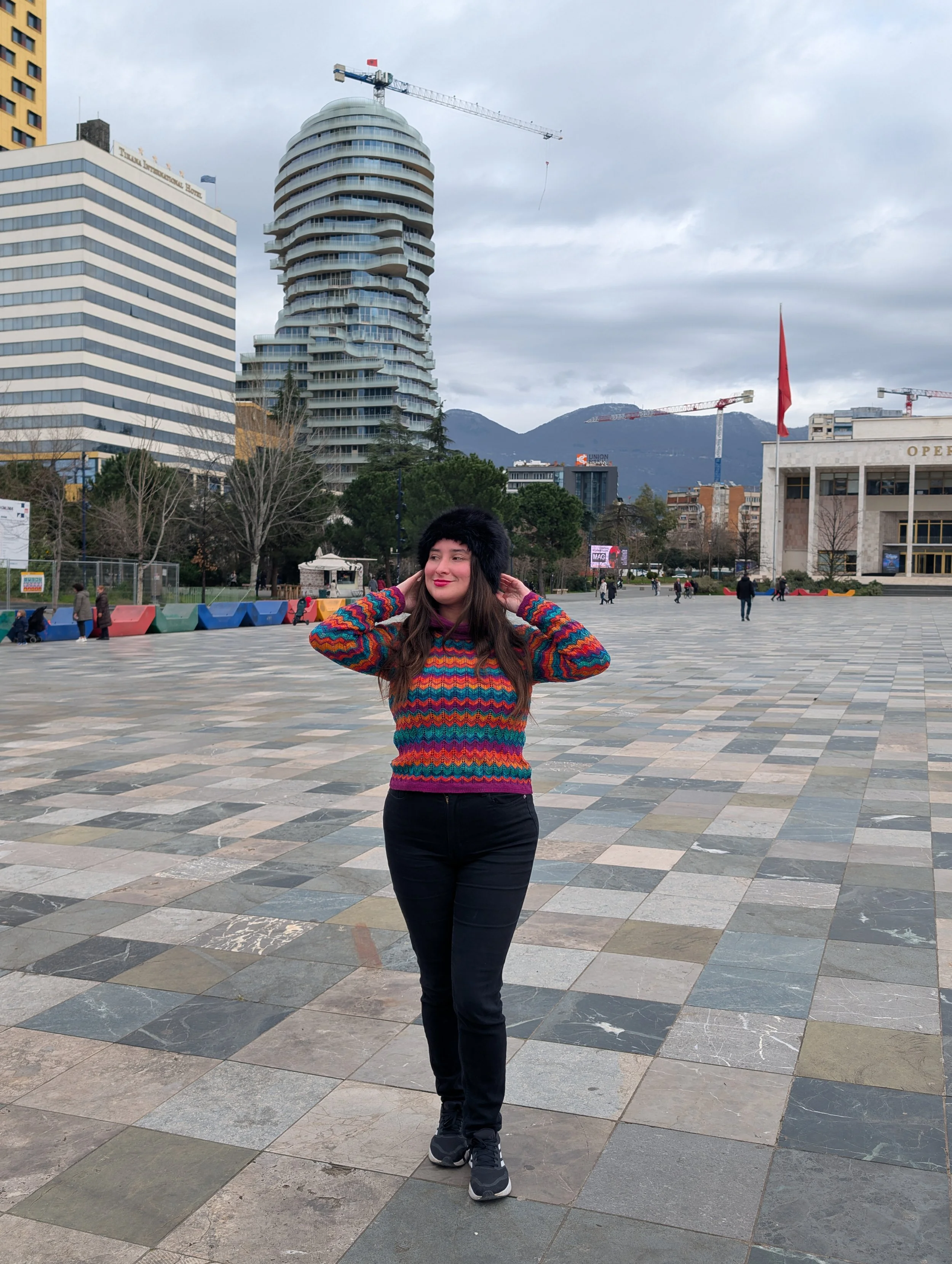 A woman in a colorful striped sweater and black pants standing on a checkered pavement in an urban area with tall buildings, trees, and construction cranes in the background.