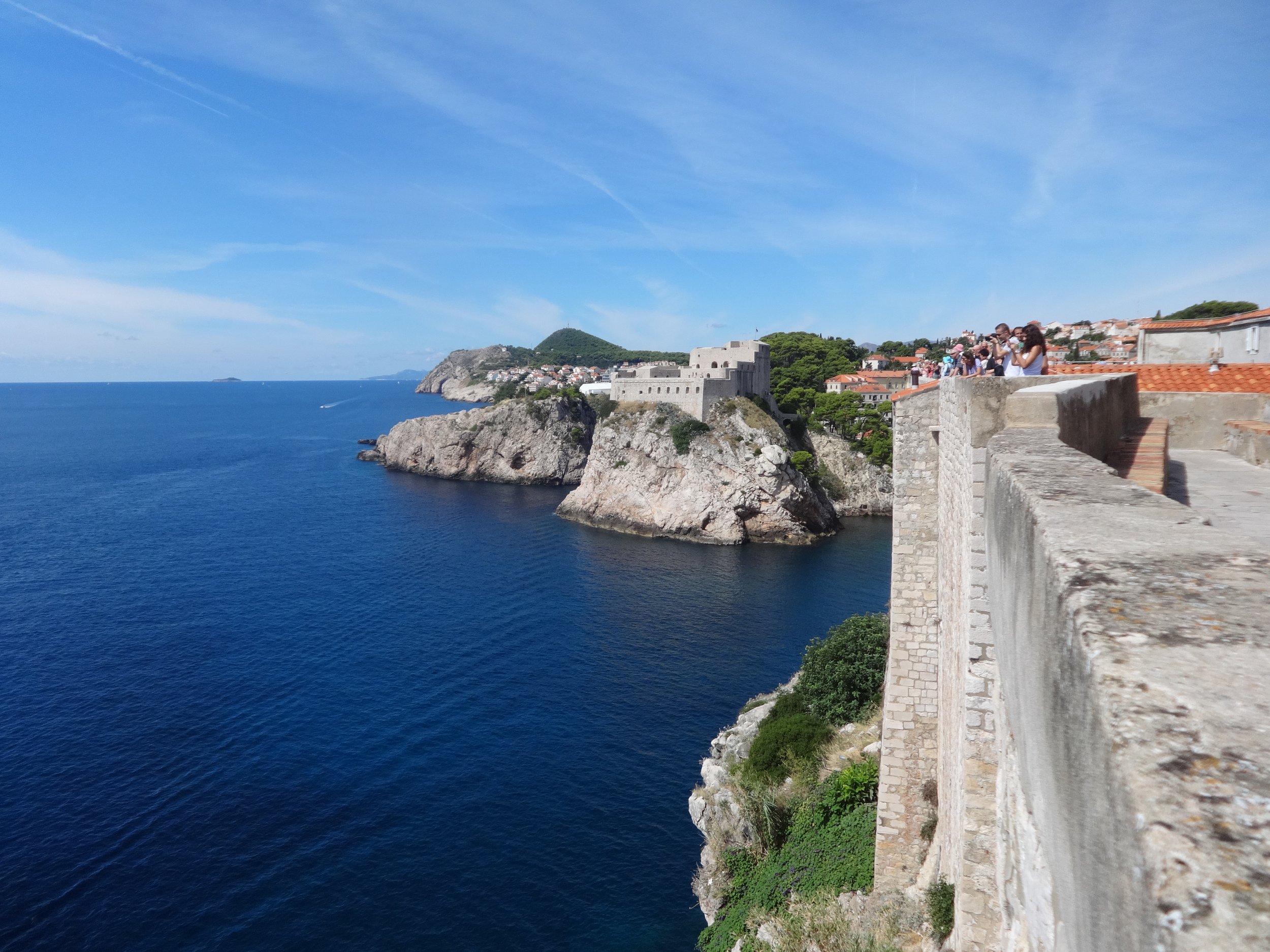 A coastal scene featuring a historic stone fortress on a rocky outcrop overlooking the deep blue sea. There is a stone wall with tourists taking photos, and a picturesque harbor town with red-tiled roofs and lush green hills in the background under a