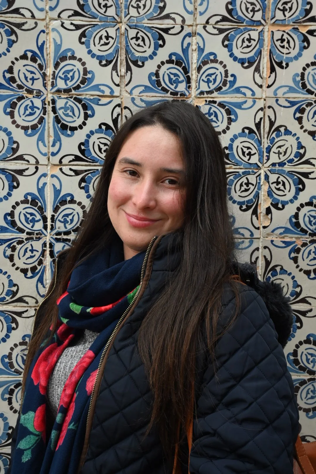 A woman with long dark hair smiling in front of a decorative blue, black, and white patterned tiled wall.
