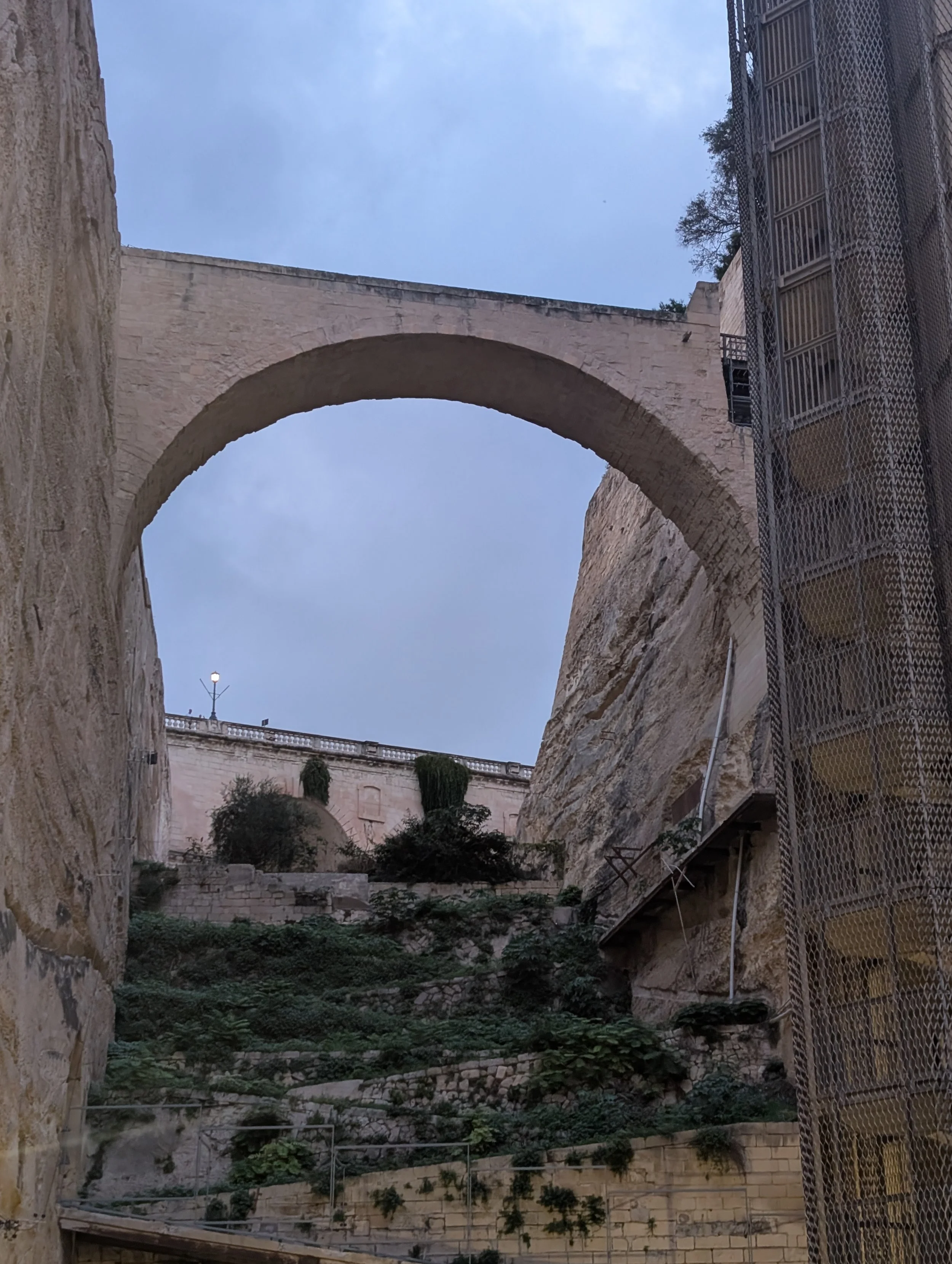 Stone arch bridge viewed from below, with buildings and trees around it, and a cloudy sky above.