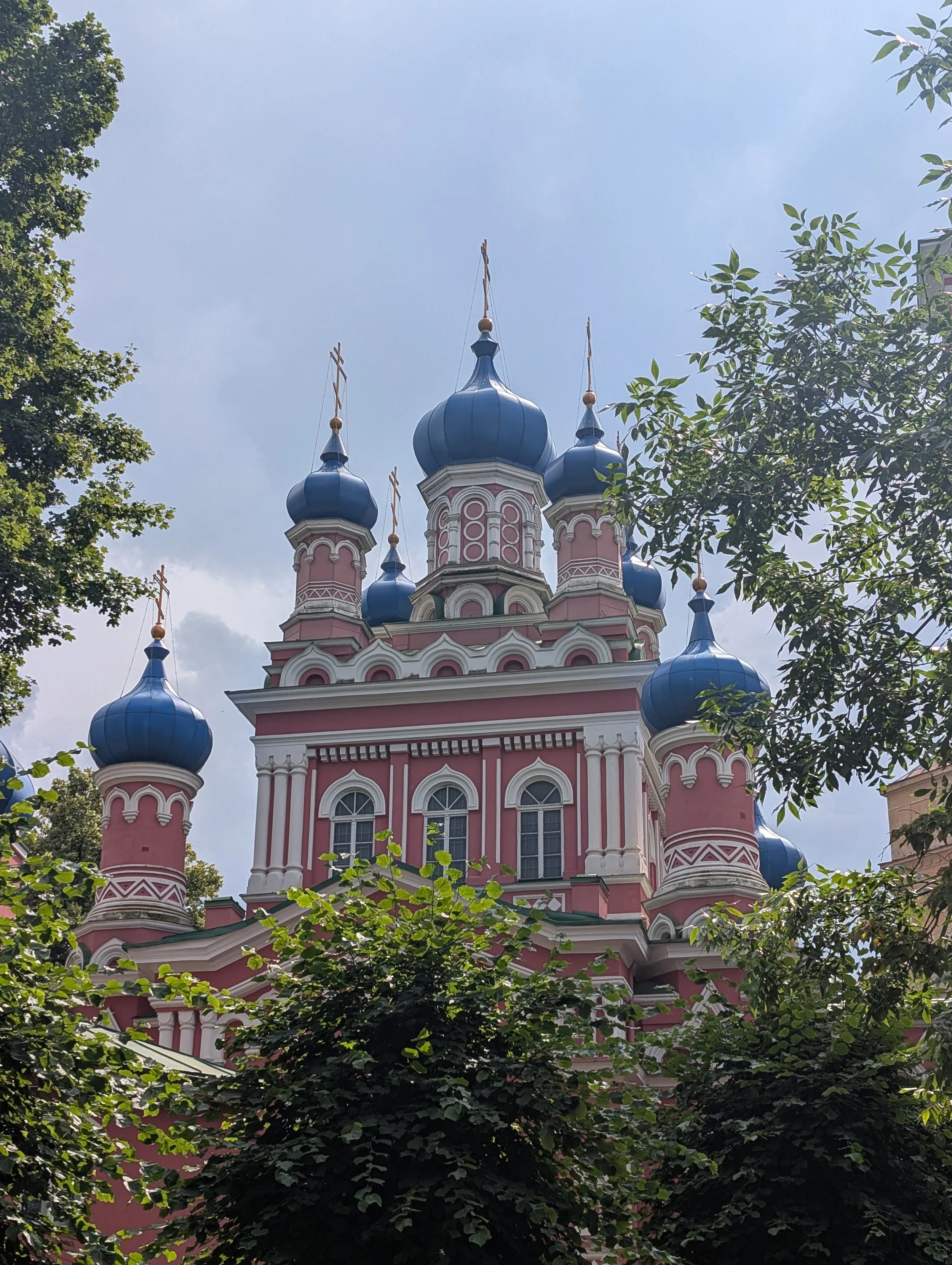 Colorful pink and white church with blue domes, surrounded by green trees, under a partly cloudy sky.