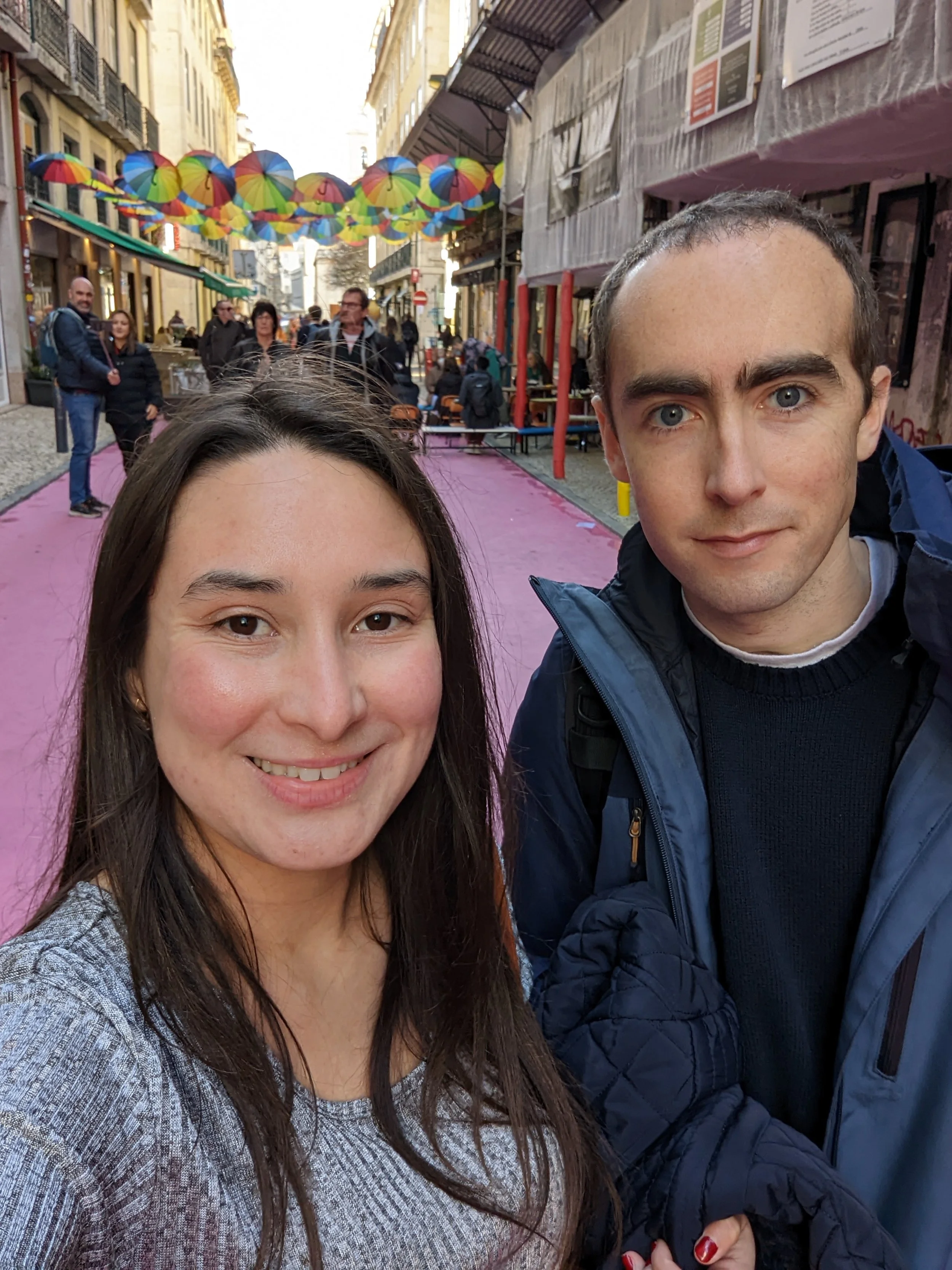 A selfie of a woman and a man in an urban street decorated with colorful umbrellas hanging above.