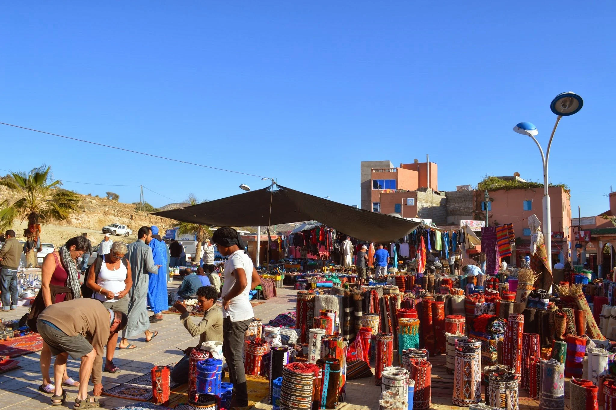 An outdoor market with various vendors selling colourful textiles, pottery, and crafts under tents. People browse and shop on a sunny day under a clear blue sky, with a hillside of buildings in the background.