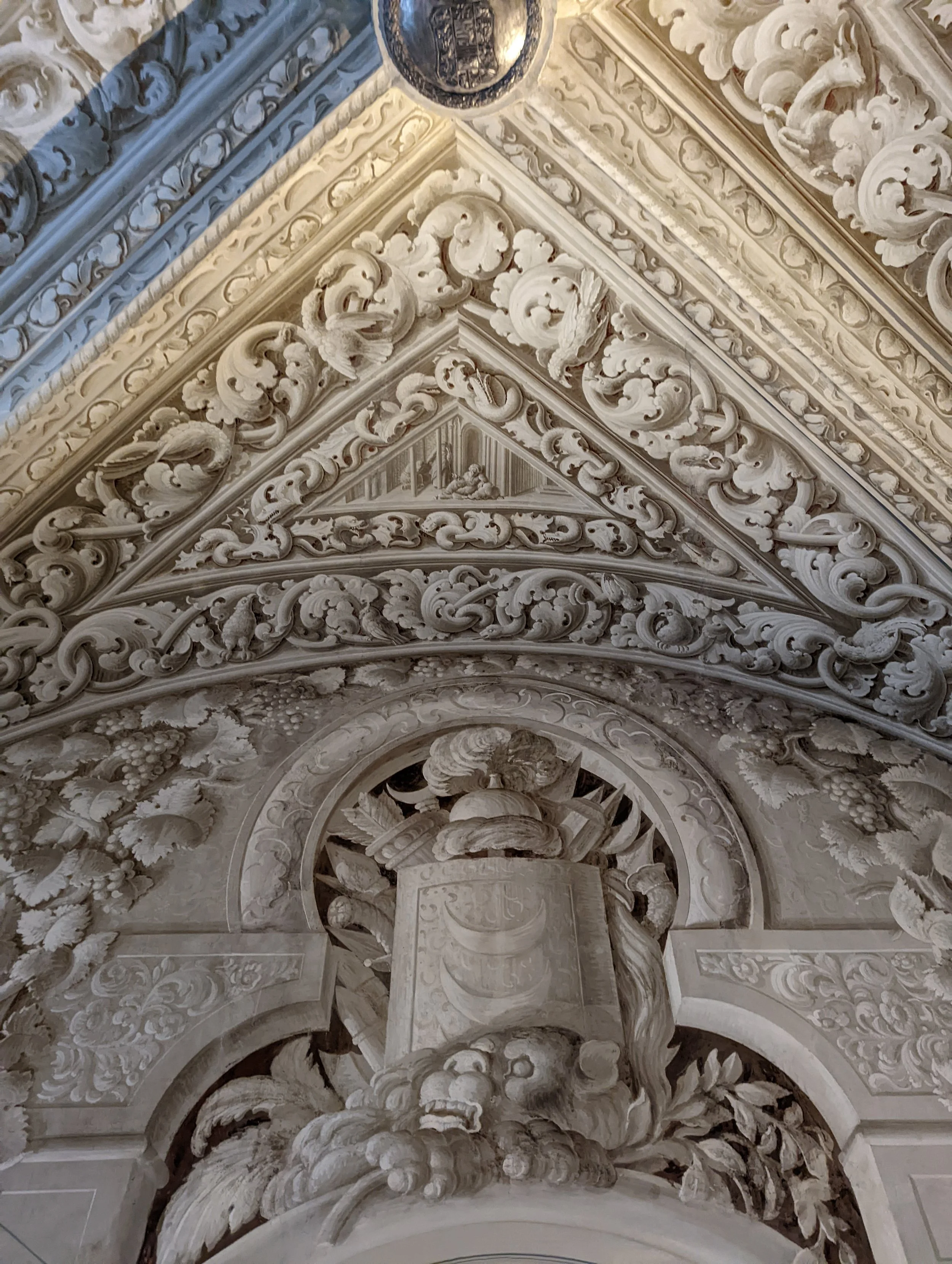 Intricate Baroque-style ceiling with elaborate stucco decorations featuring swirling patterns, floral motifs, cherub figures, and a central sculpture.