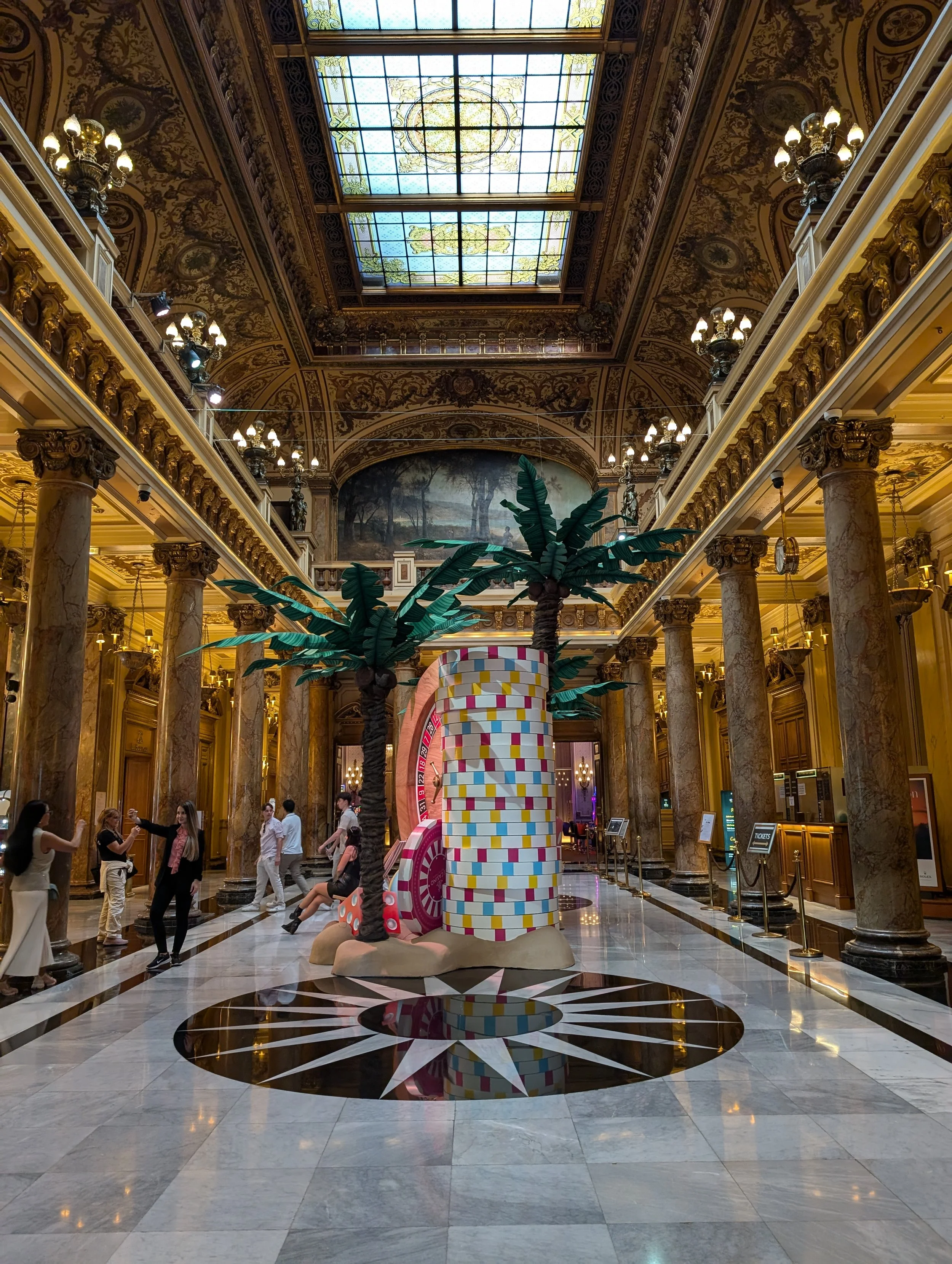 Interior of a grand, ornate building with high ceilings, stained glass skylights, marble columns, and a decorative floor. There is a large, colorful, abstract sculpture in the center with two artificial palm trees. Several people are walking and taki