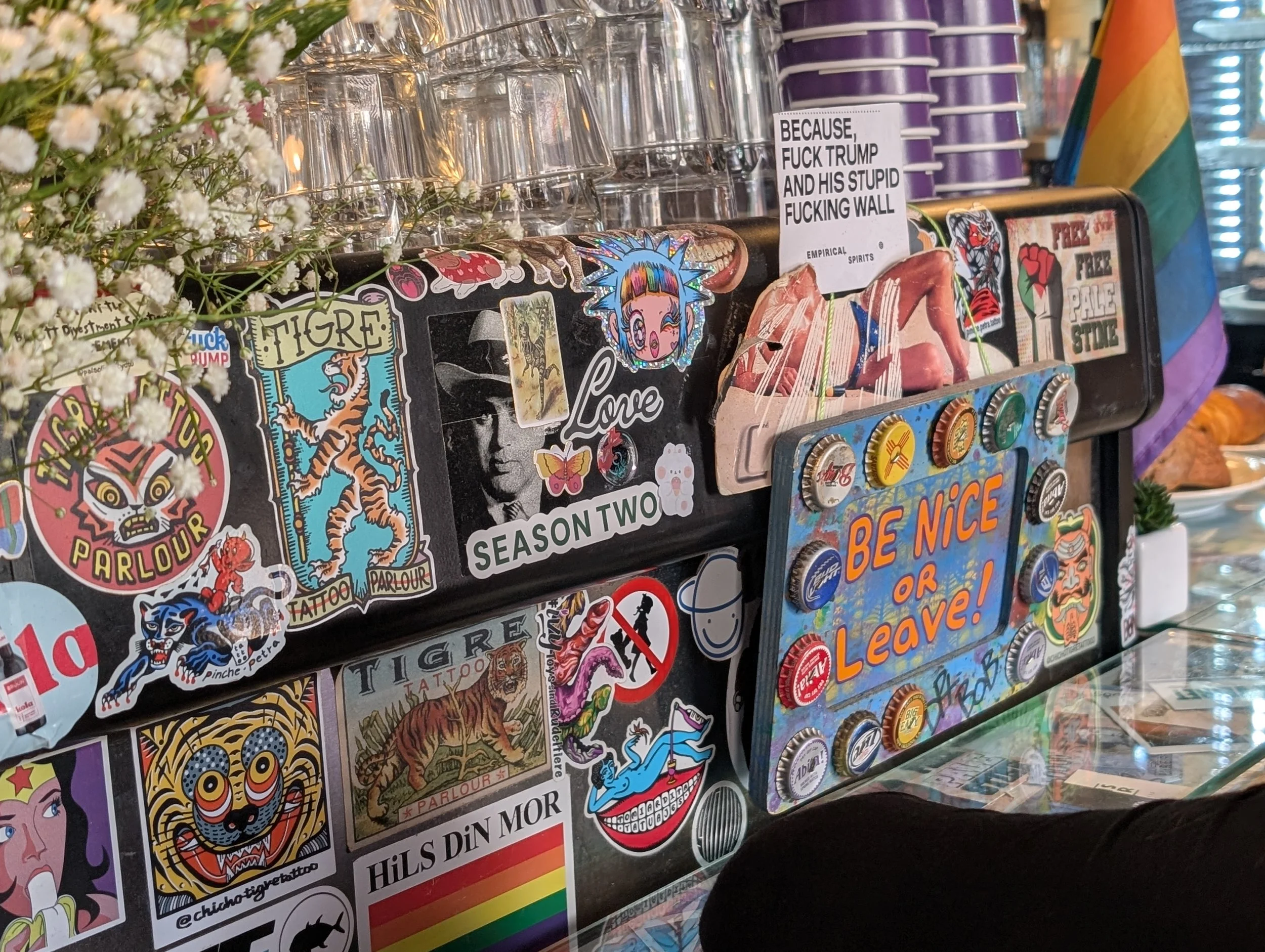 A display of various colorful stickers on a surface, with some greenery on the left and rainbow-colored pride flags on the right. The stickers include images of skulls, tigers, and cartoon characters, and some have text such as "BE NICE OR LEAVE" and