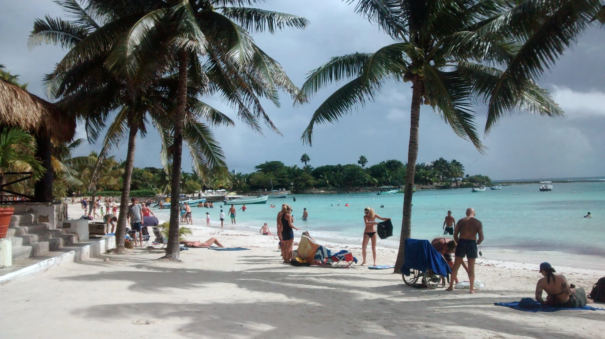People enjoying a sunny day at a tropical beach with white sand, palm trees, and calm turquoise water. Several boats are anchored near the shore, and many people are swimming, sunbathing, and walking along the beach.