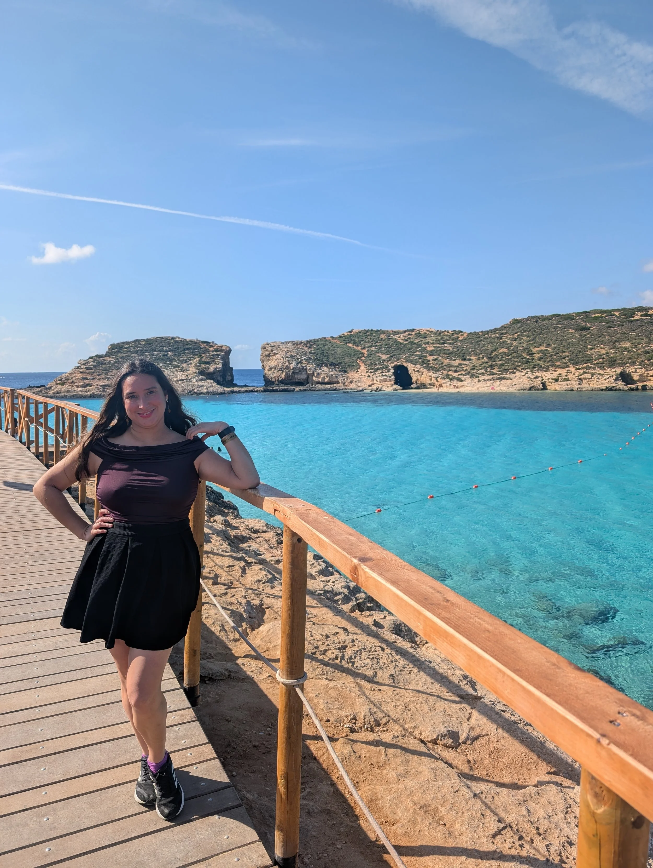 A woman standing on a wooden walkway by the beach, with rocky cliffs and a cave in the background, and clear blue water.