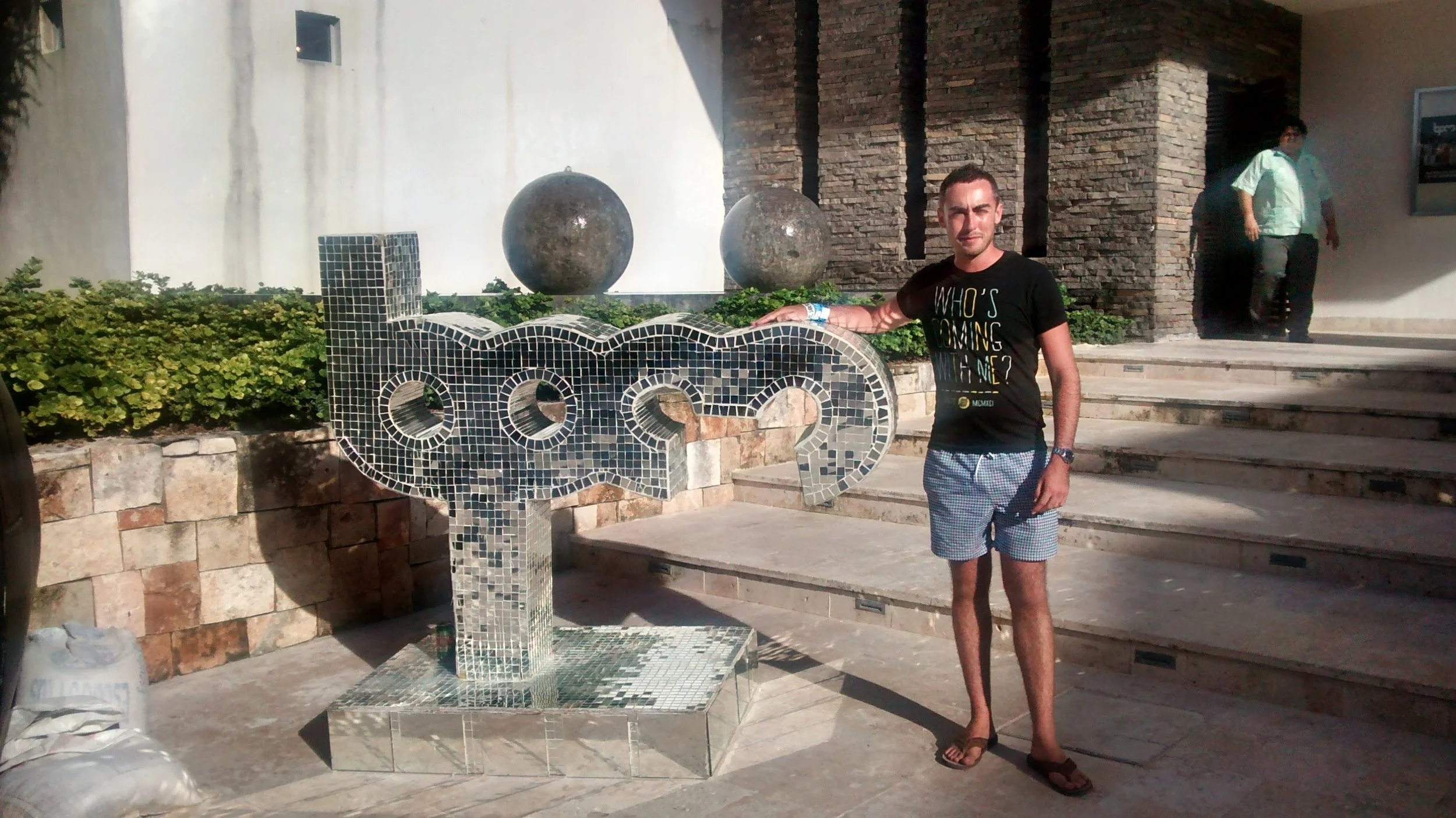 A man stands next to a mosaic sculpture spelling 'LOVE' outdoors, with green plants and stone stairs in the background.