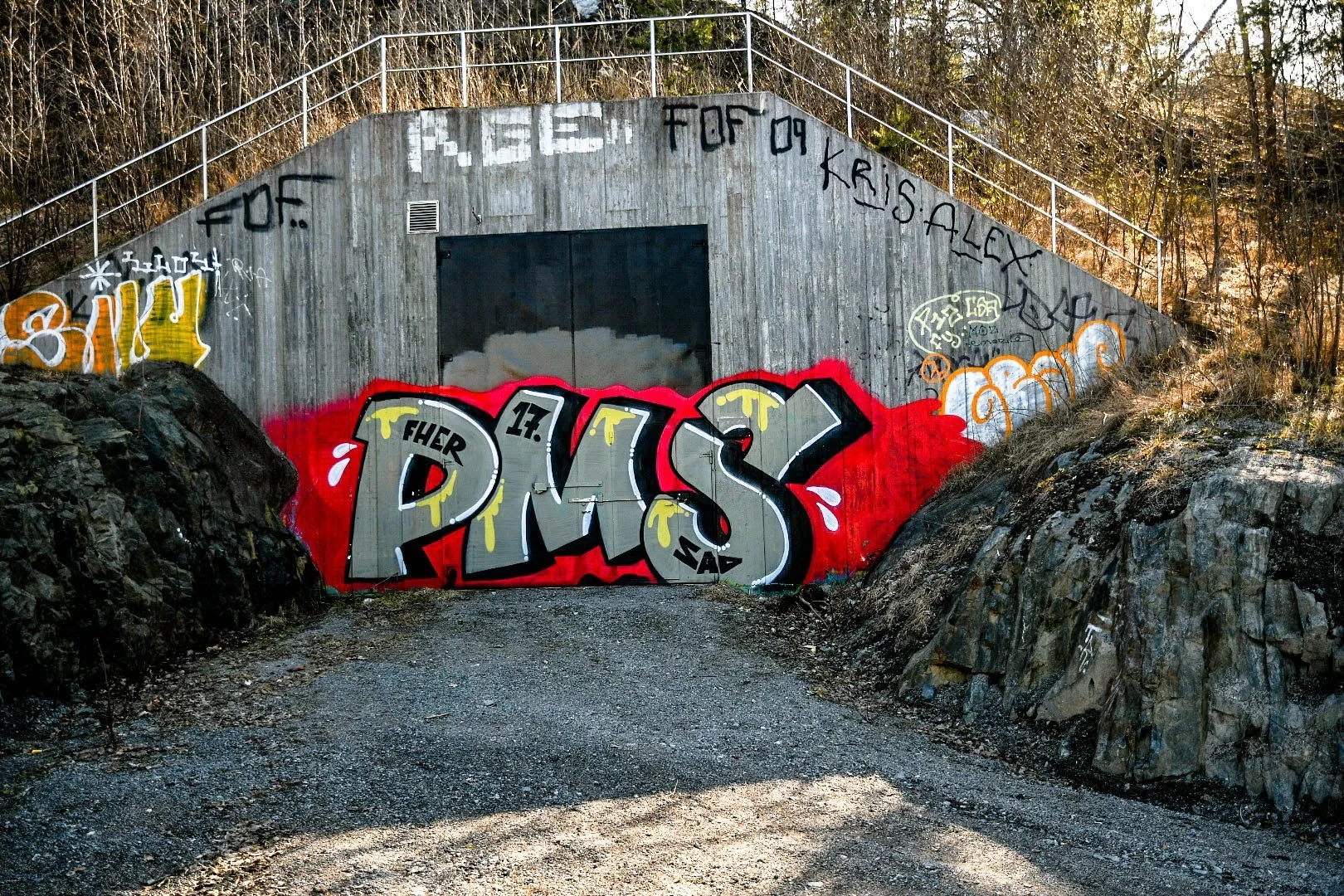 Graffiti art on a concrete structure situated between rocky terrain and a wooded background. The graffiti features colorful, bold letters spelling out "PRS" with additional smaller tags and phrases around it.
