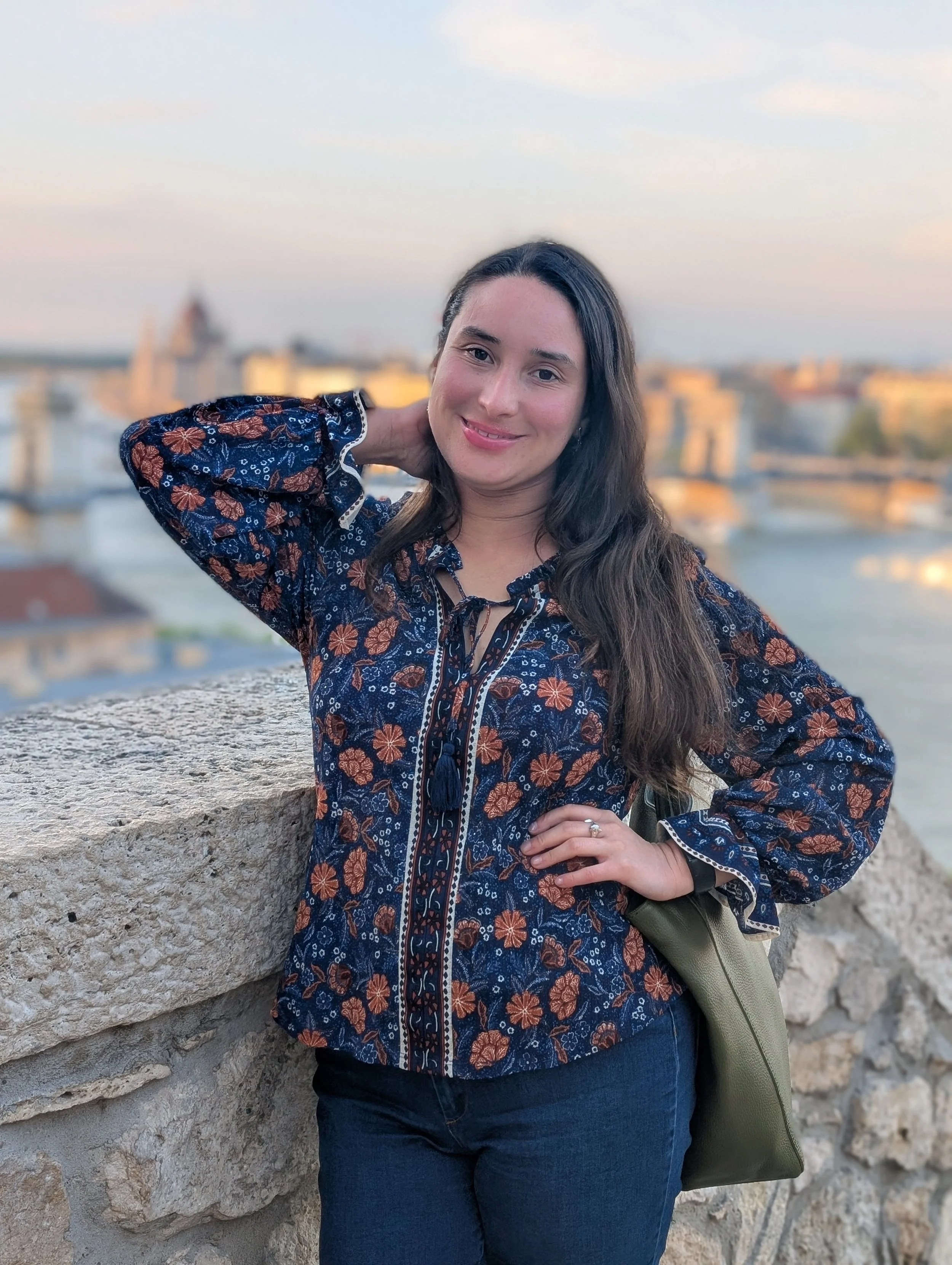 A woman with long dark hair and a floral patterned blouse posing outdoors by a stone wall with a cityscape and a river in the background at sunset.