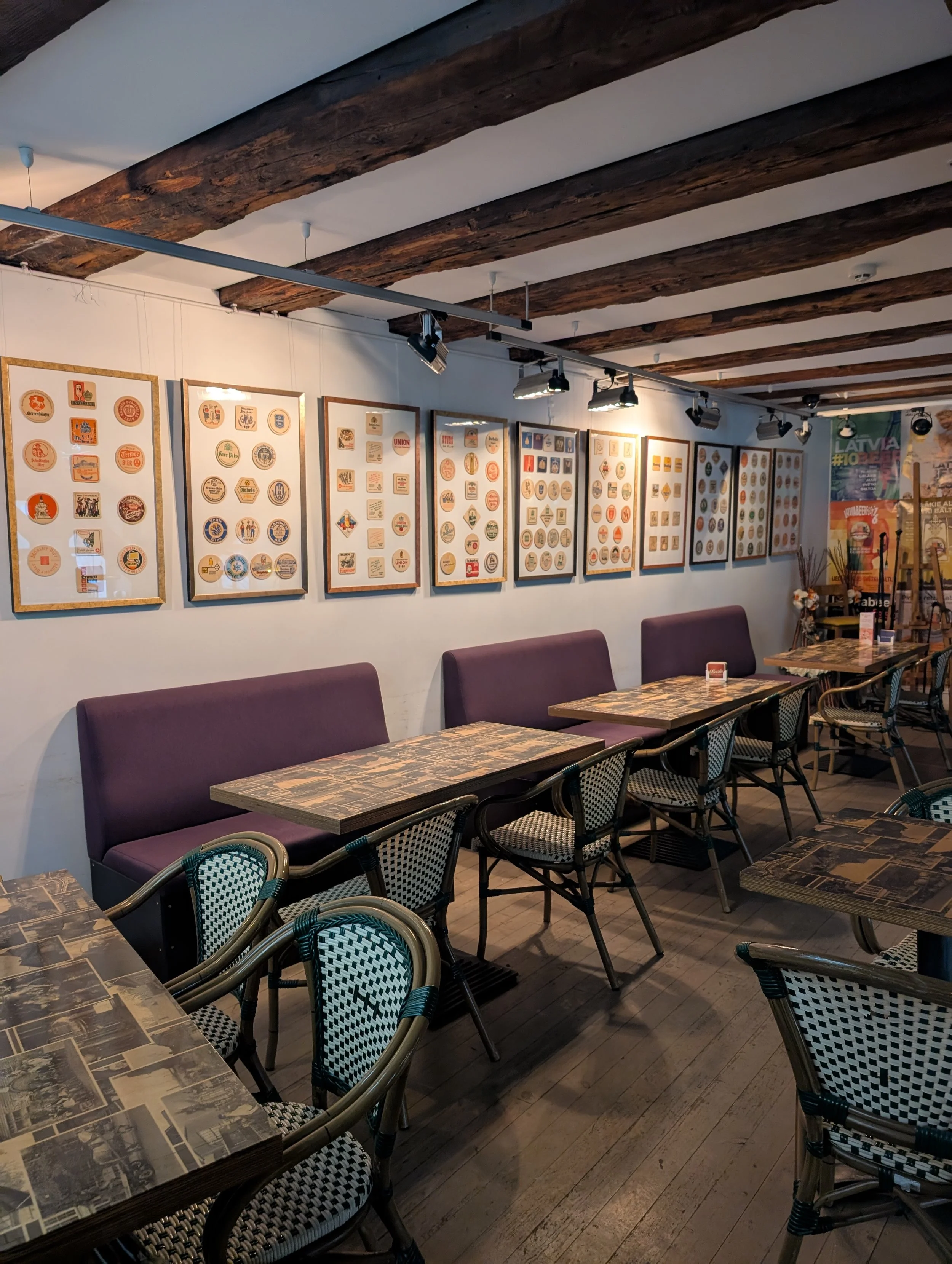 Inside a restaurant or cafe with empty tables and chairs, framed artwork on the white wall, exposed wooden ceiling beams, and dim lighting.