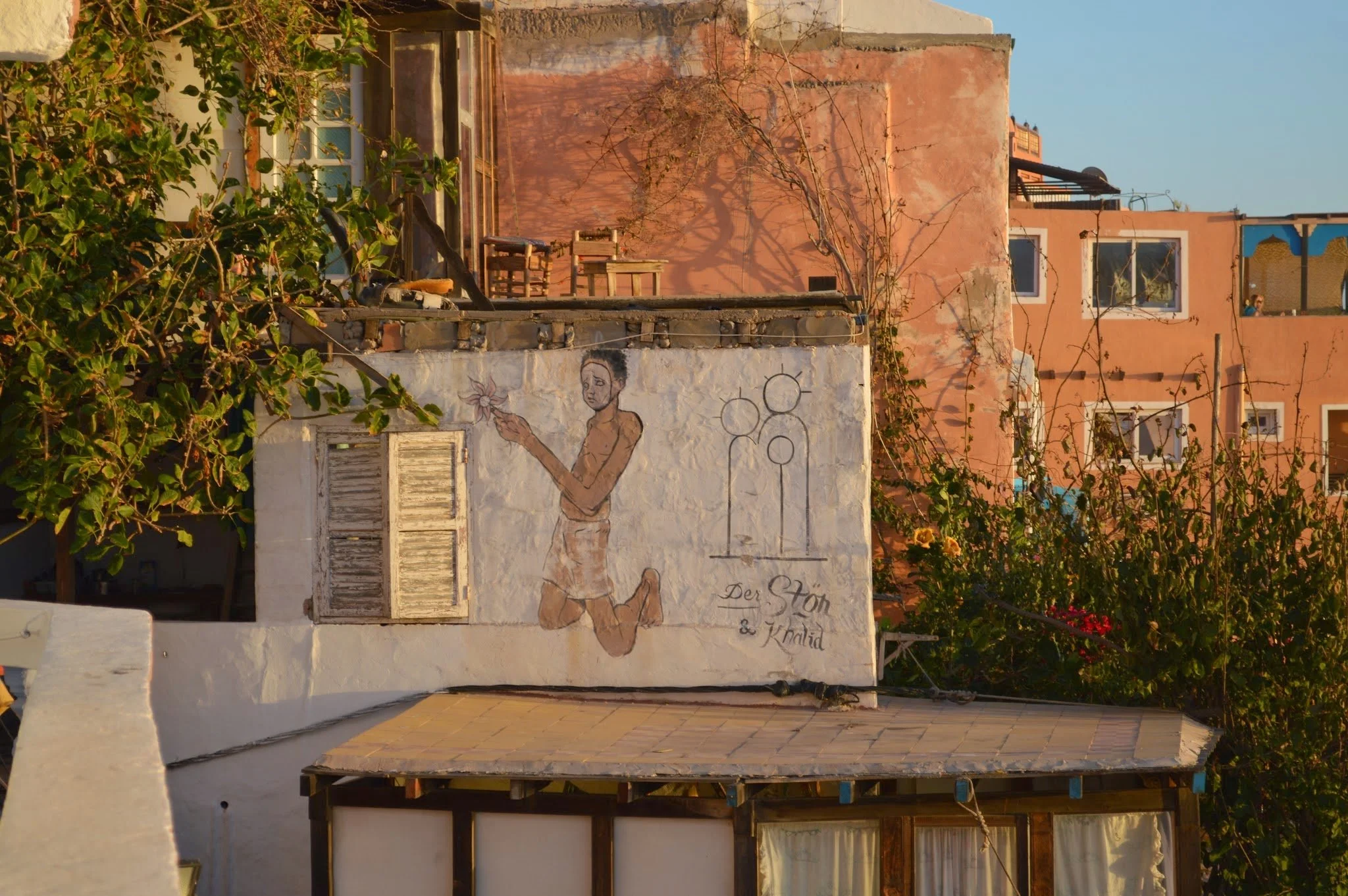 A white wall with a mural of a woman holding a flower and the phrases 'Der Stolz &' and 'Knald' written on it. There is a small window with closed shutters, and a tree branch casting shadows on the wall. Part of a building with pinkish-orange walls a