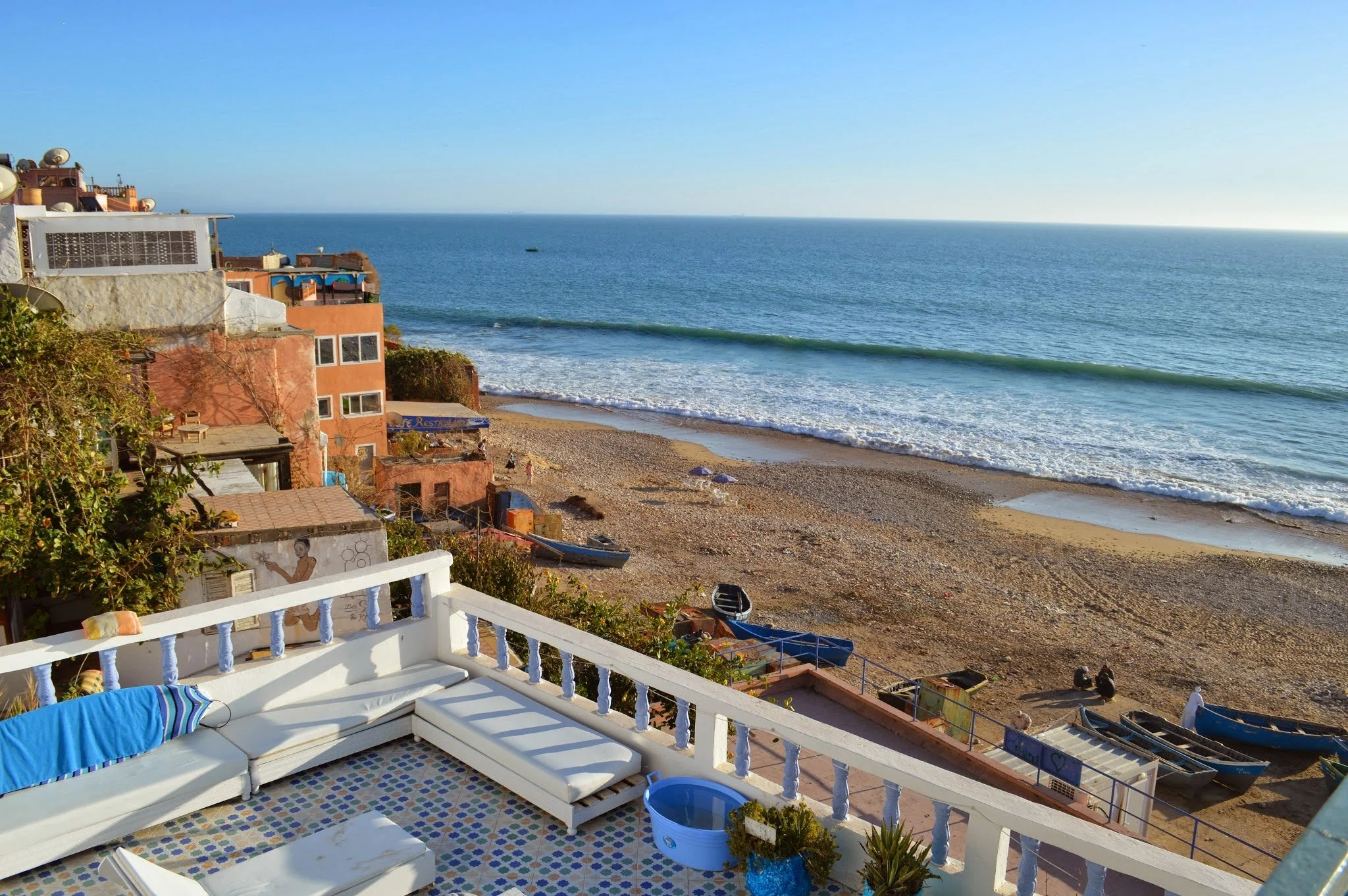 Balcony with white railing and colourful tiled floor overlooking a beach with houses and the ocean in the background.