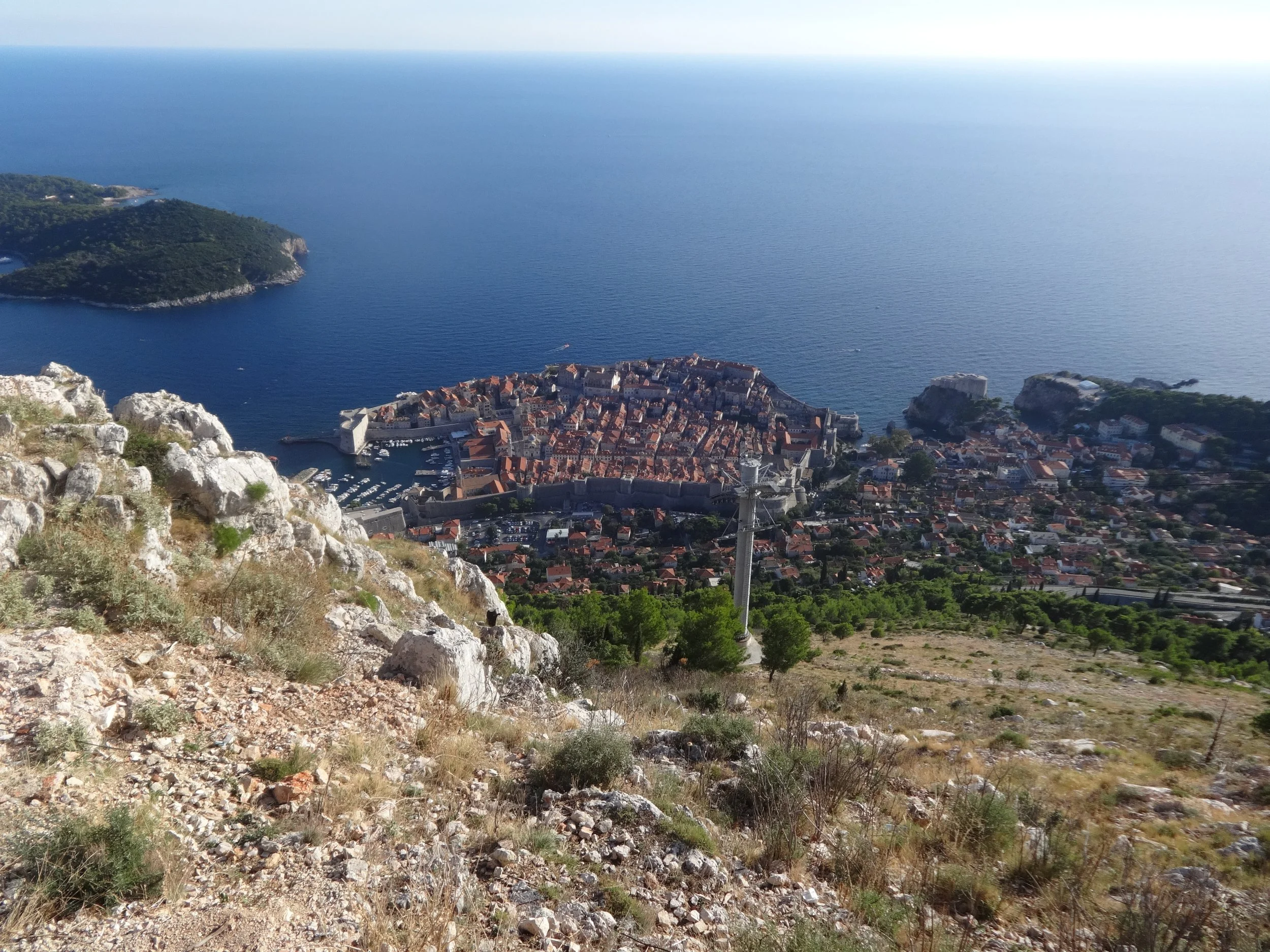 View from a hilltop overlooking a historic coastal city with a harbor, old city walls, red-tiled roofs, and the sea in the background.